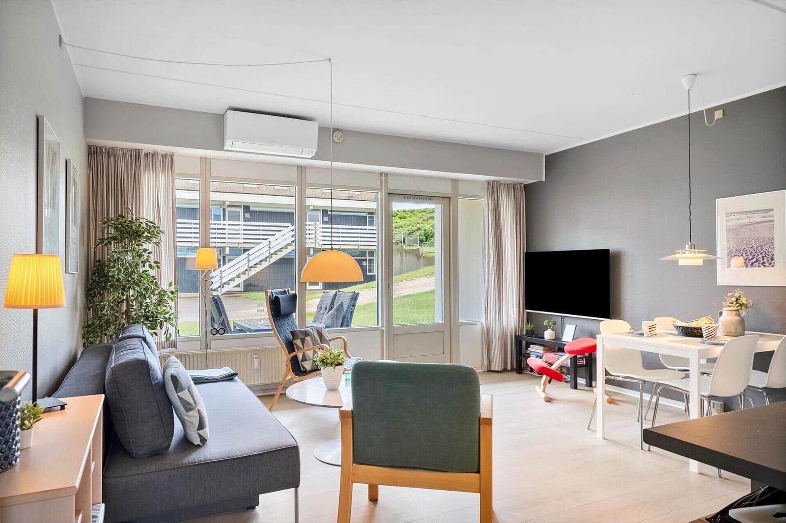 Wohn- und Essbereich mit Sofa, Stühlen und TV. Große Fenster mit Blick auf Außenbereich.