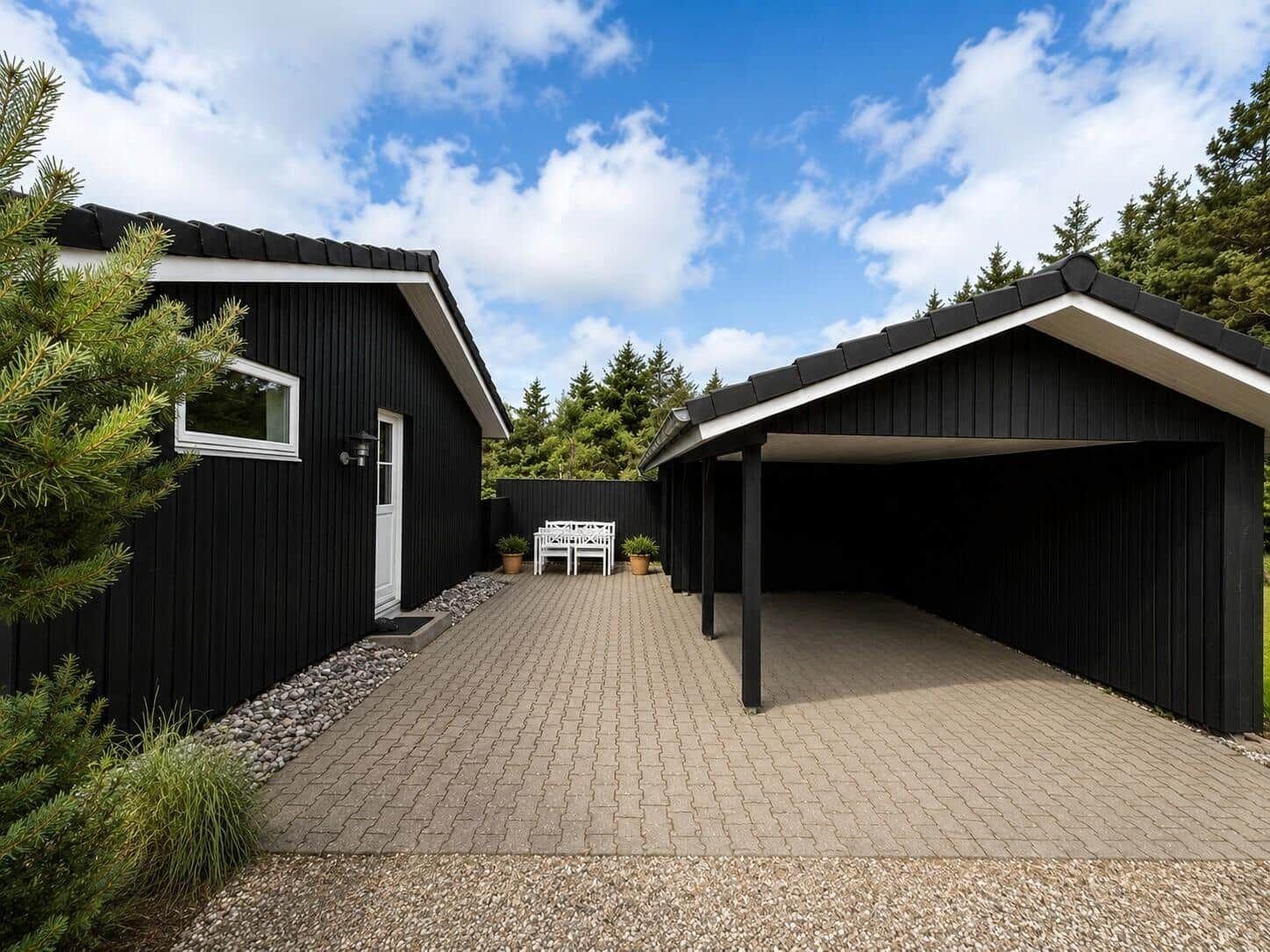 Schwarze Holzhütte mit Carport und gepflastertem Hof. Weiße Bank und Pflanzen im Garten.