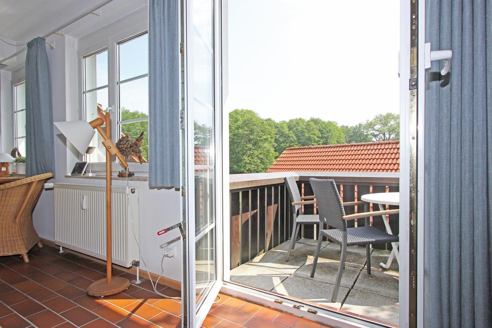 Terrasse mit Tisch und Stühlen, Blick auf Dach und Bäume. Fenster öffnet sich in Wohnraum mit Holzfußboden.