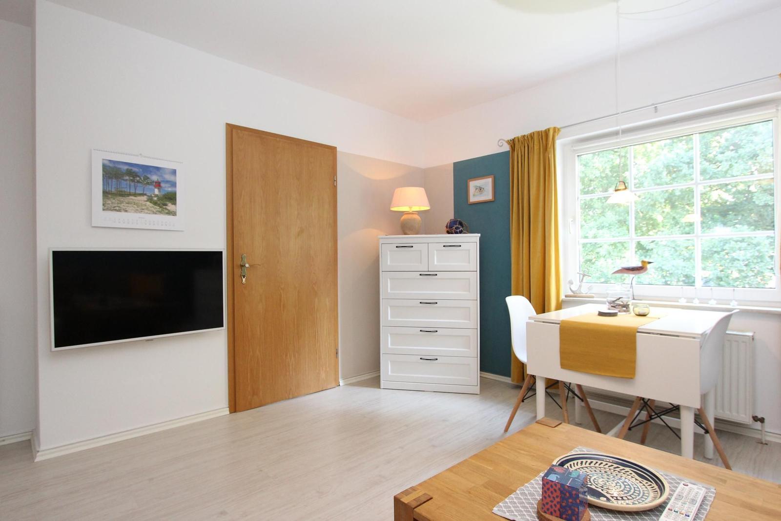 Ein Raum mit Fernseher, Schrank, Tisch und Fenster mit gelben Vorhängen.