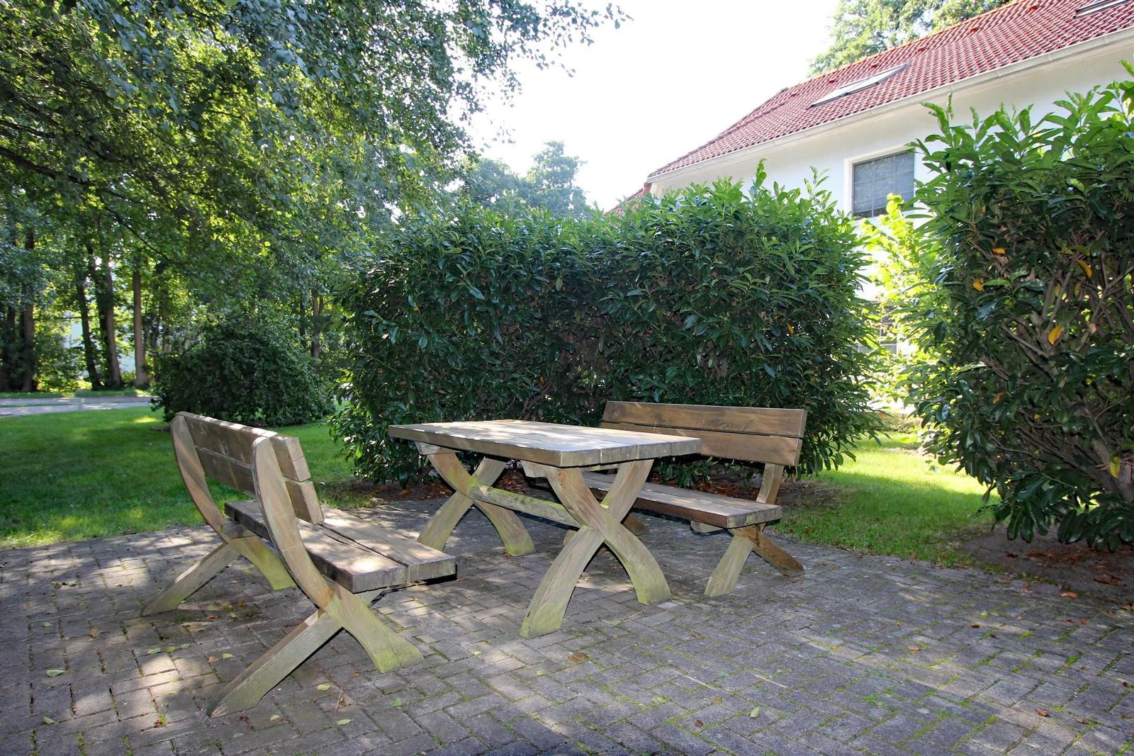 Holztisch und Bänke auf gepflastertem Platz vor grüner Hecke und Haus