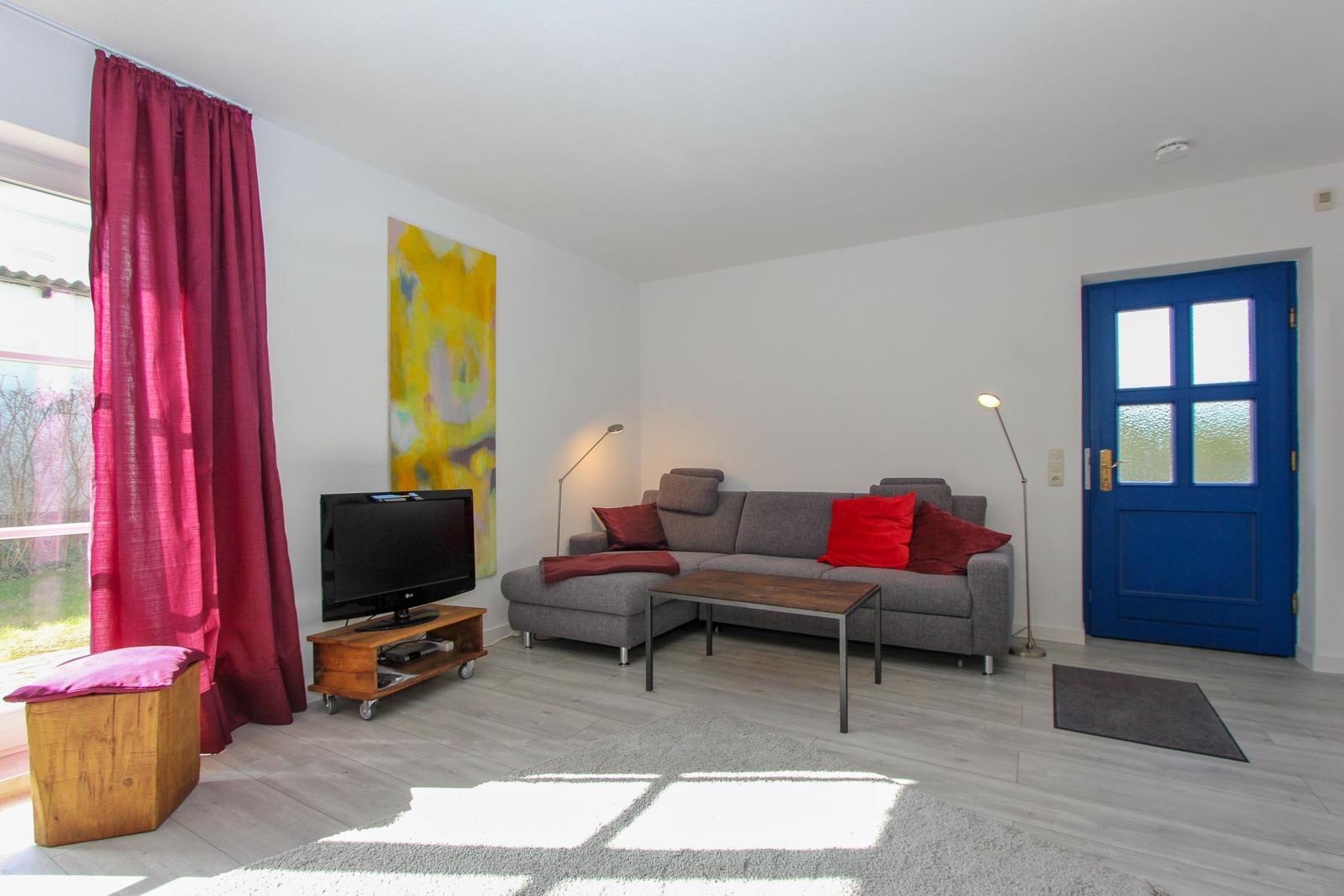 Wohnzimmer mit grauem Sofa, roten Kissen, blauer Tür und Fernseher auf Holzstand.