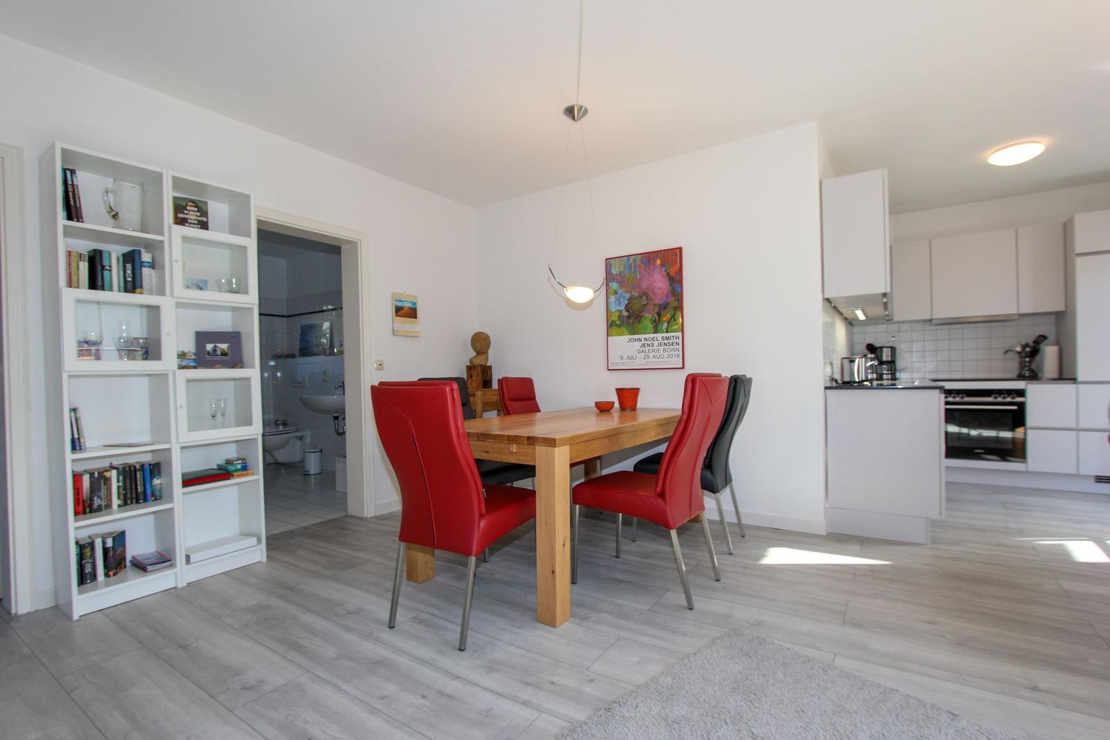 Essbereich mit Holztisch, roten Stühlen und weißen Küchenmöbeln. Bücherregal und Wandbild sichtbar.