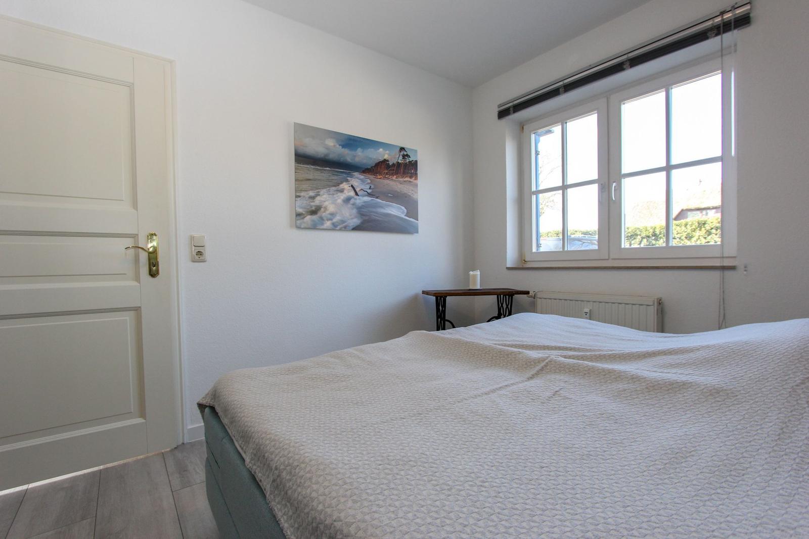 Schlafzimmer mit Bett, Fenster und Wandbild. Tür links, Tisch rechts.