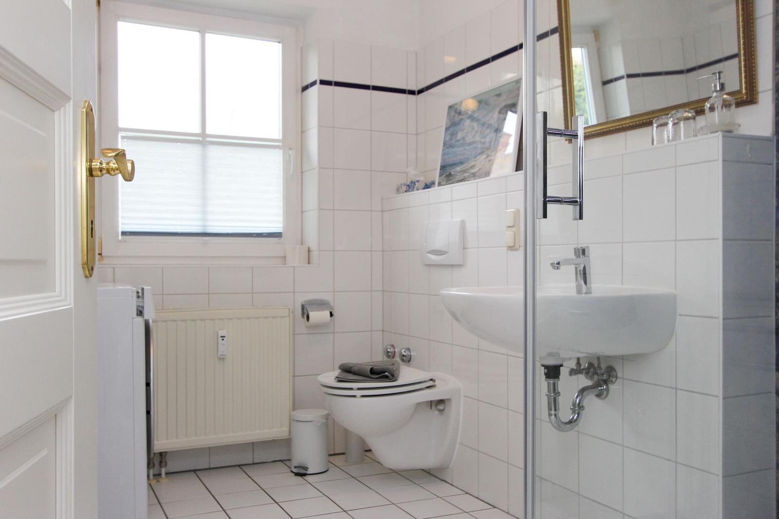 Badezimmer mit Toilette, Waschbecken und Dusche. Weiße Fliesen und Fenster mit Jalousie.