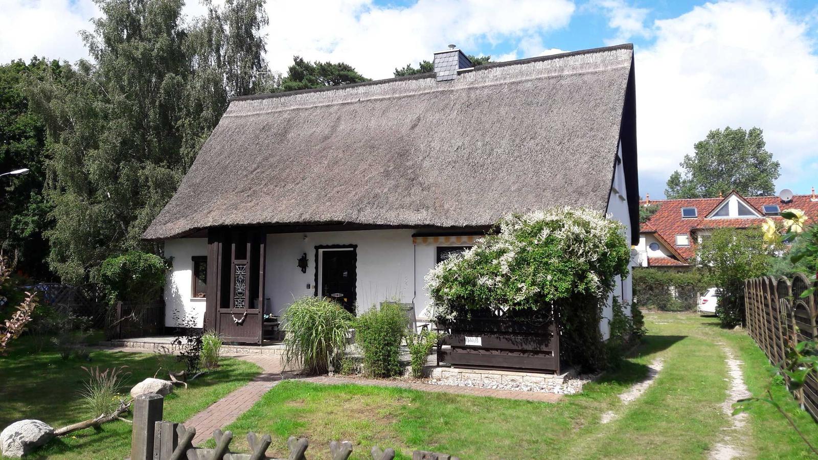 Ein Haus mit Strohdach, weißer Fassade und Garten. Holztor und Pflanzen sind sichtbar.