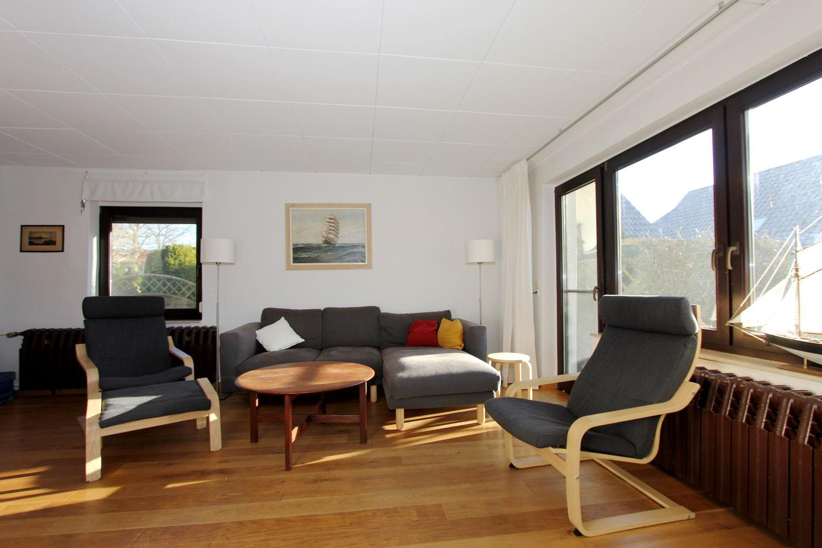 Wohnzimmer mit Sofa, zwei Sesseln, Tisch und großen Fenstern.