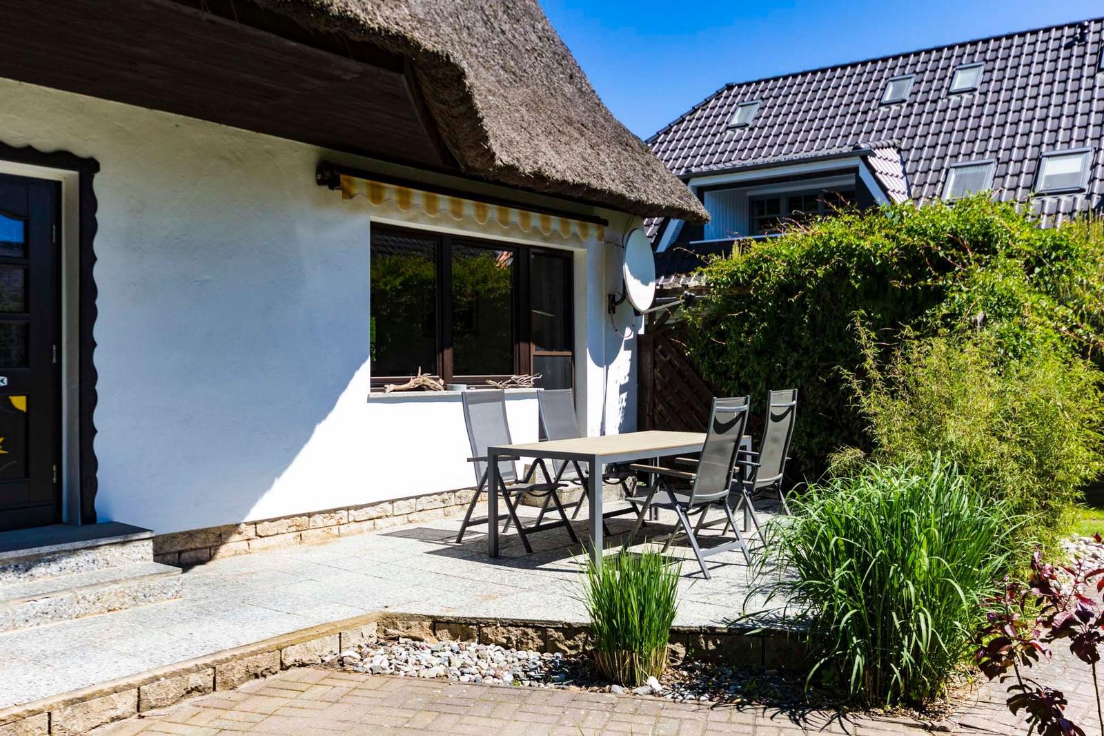 Haus mit Dach und Terrasse mit Tisch und Stühlen. Hinterhof mit Hecke und Pflanzen.