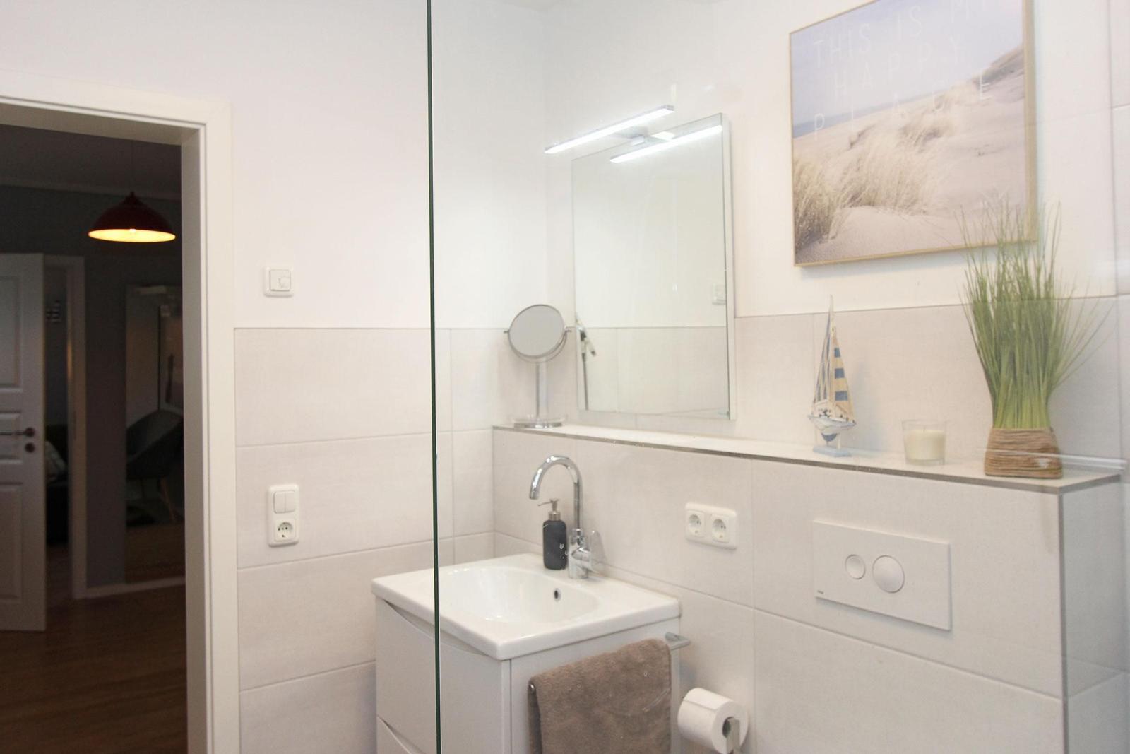 Badezimmer mit Spiegel, Waschbecken und Glaswand. Dekoration mit Bild und Pflanze.