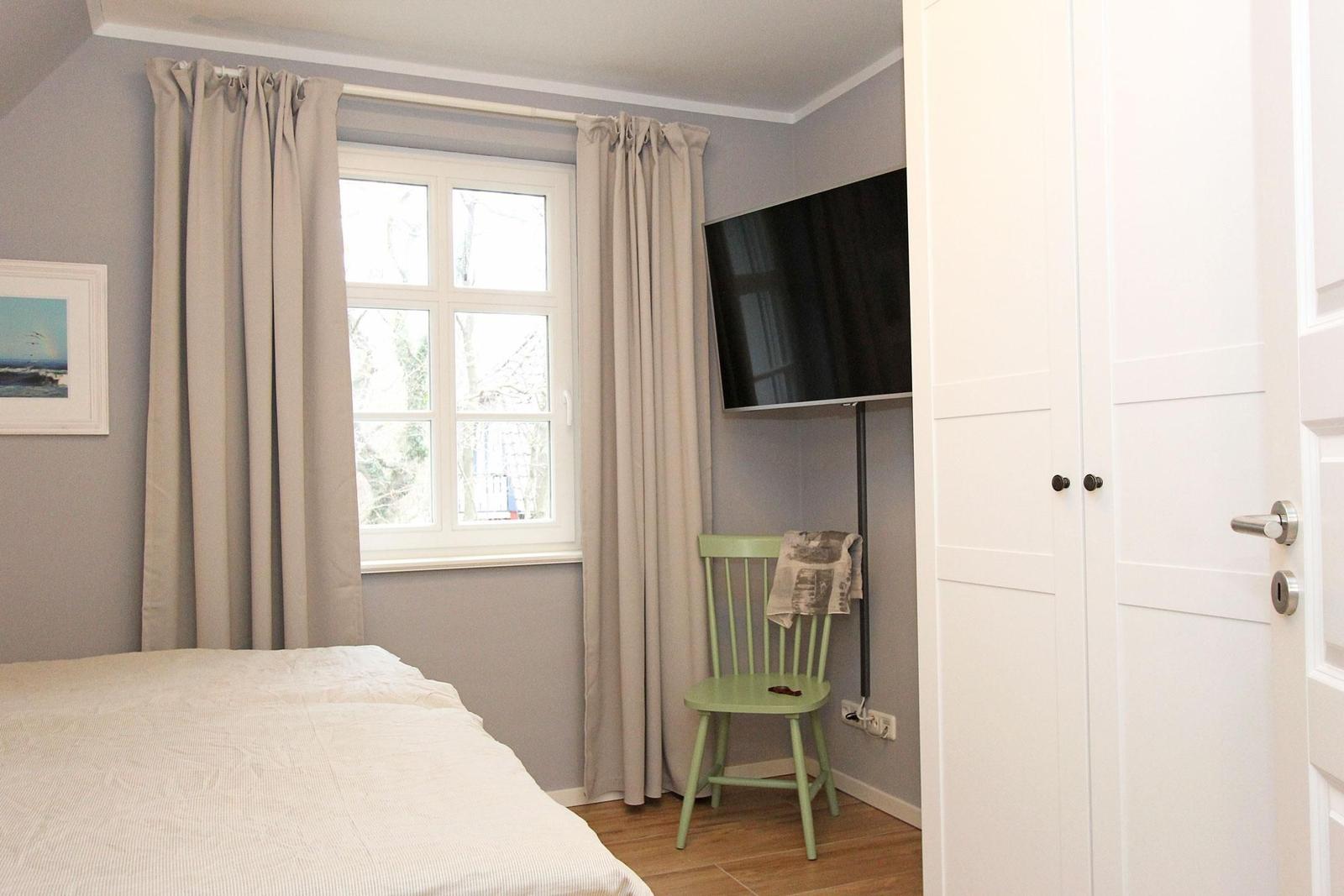 Schlafzimmer mit Bett, Fenster, TV und grüner Stuhl.