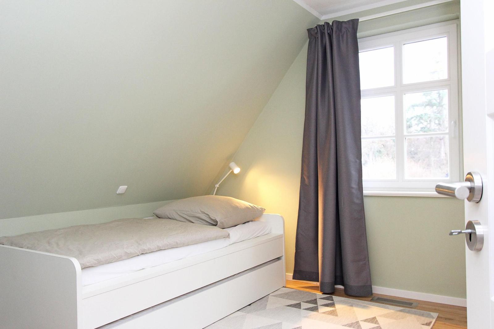 Ein Schlafzimmer mit Dachschrägen, weißem Bett, grauem Vorhang und Fenster.