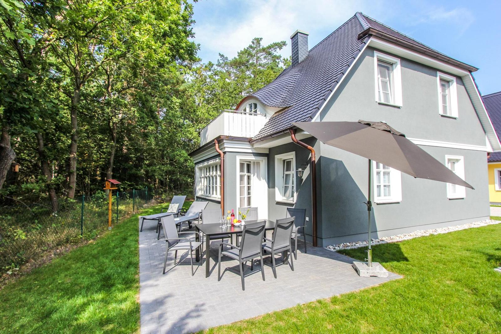 Graues Haus mit Terrasse, Tisch, Stühlen und Sonnenschirm. Umgeben von Grün und Bäumen.