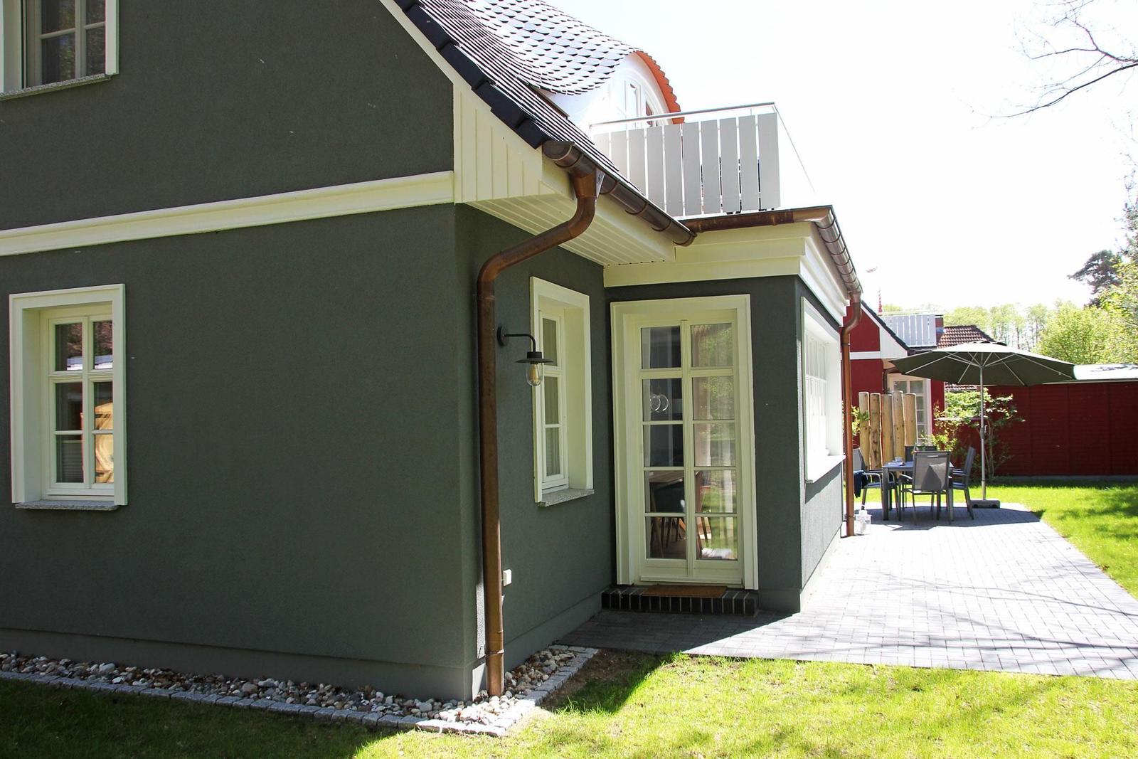Ein Haus mit grauer Fassade, weißer Tür und Balkon. Außenbereich mit Tisch, Stühlen und Sonnenschirm.
