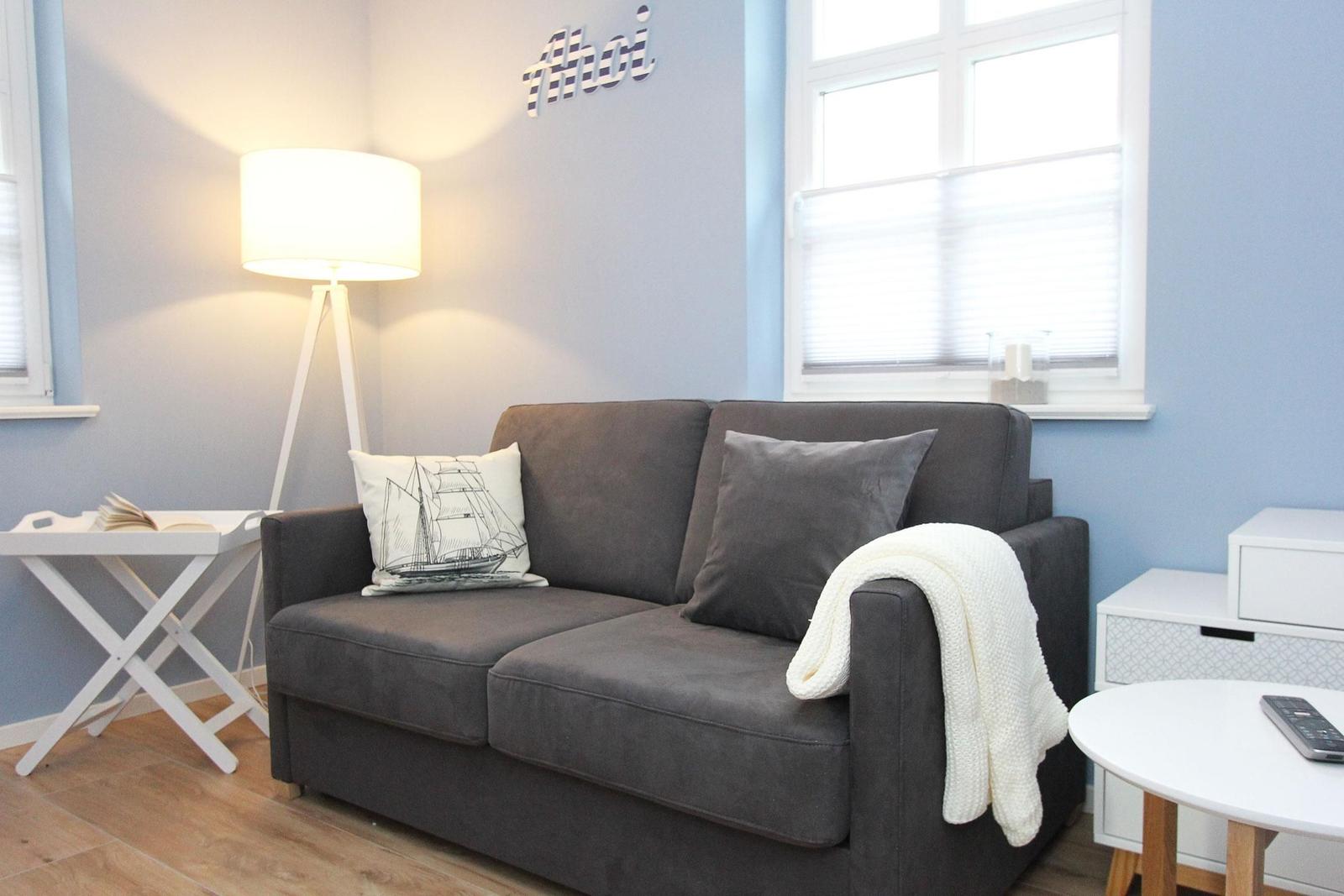 Ein grauer Sofa mit Kissen und Decke, Wand mit Schriftzug, Fenster und Beleuchtung.