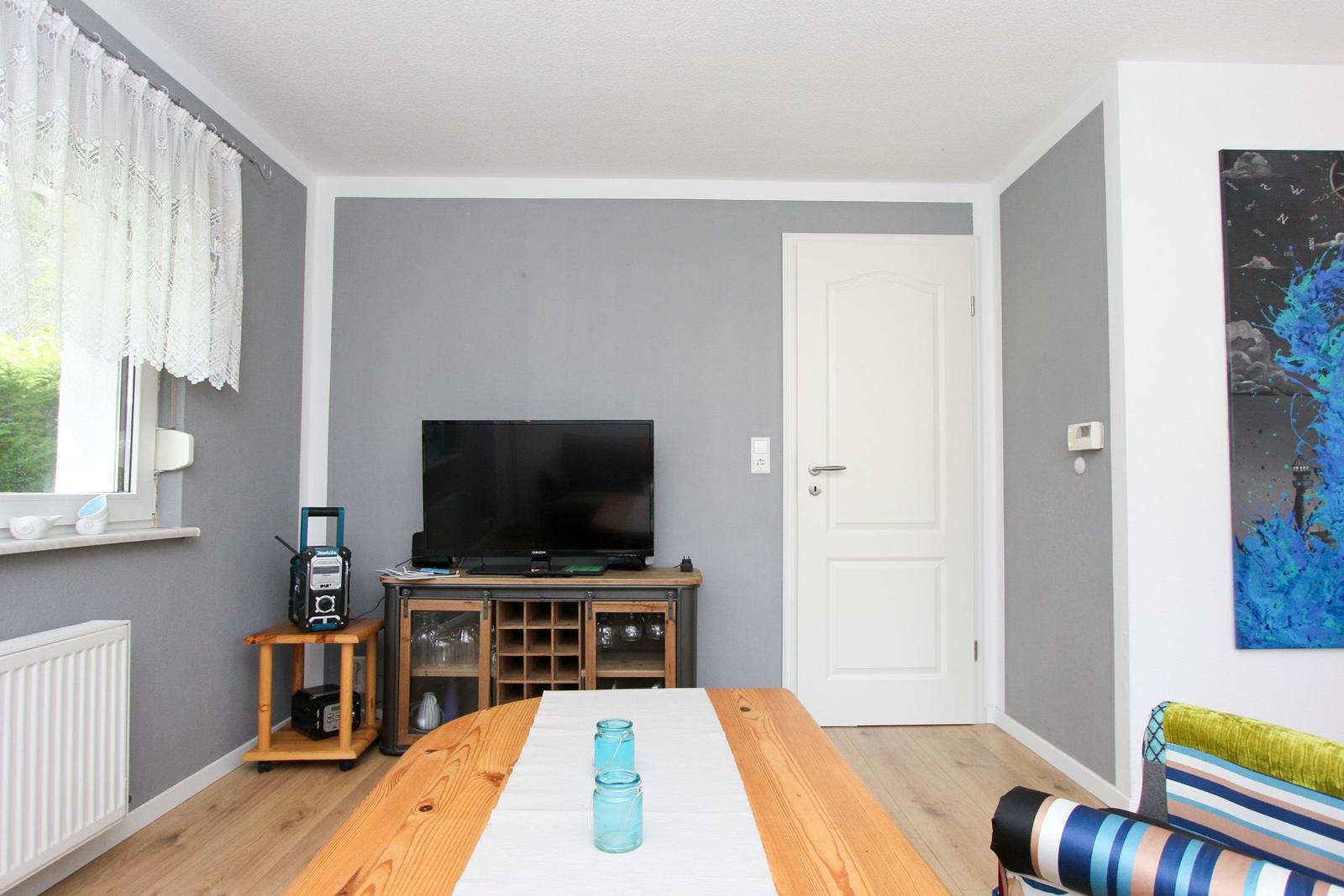 Ein Raum mit Holztisch, Fernseher, Tür und Fenster mit weißen Vorhängen.