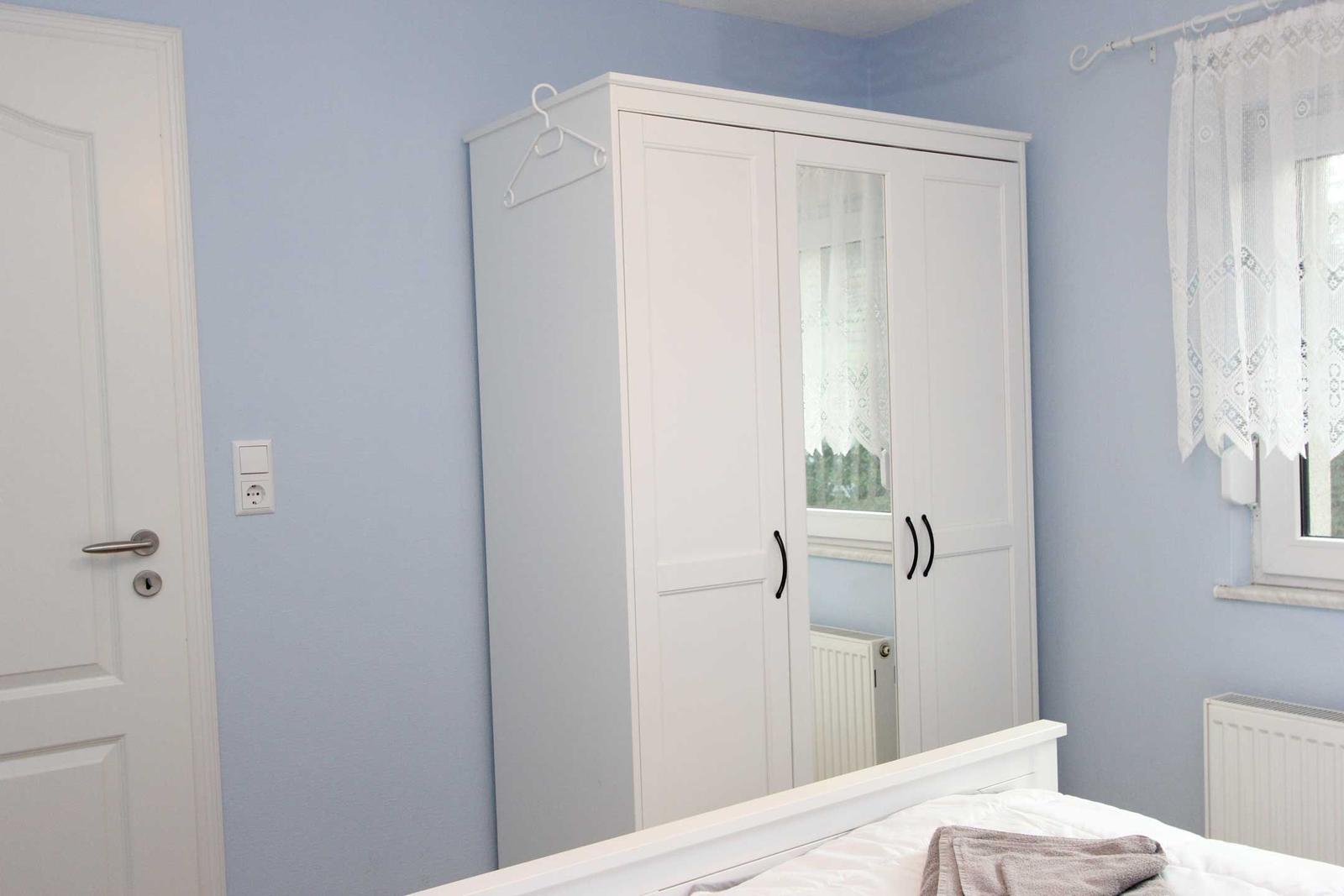 Schlafzimmer mit weißem Kleiderschrank, Bett und Fenster mit Vorhang.