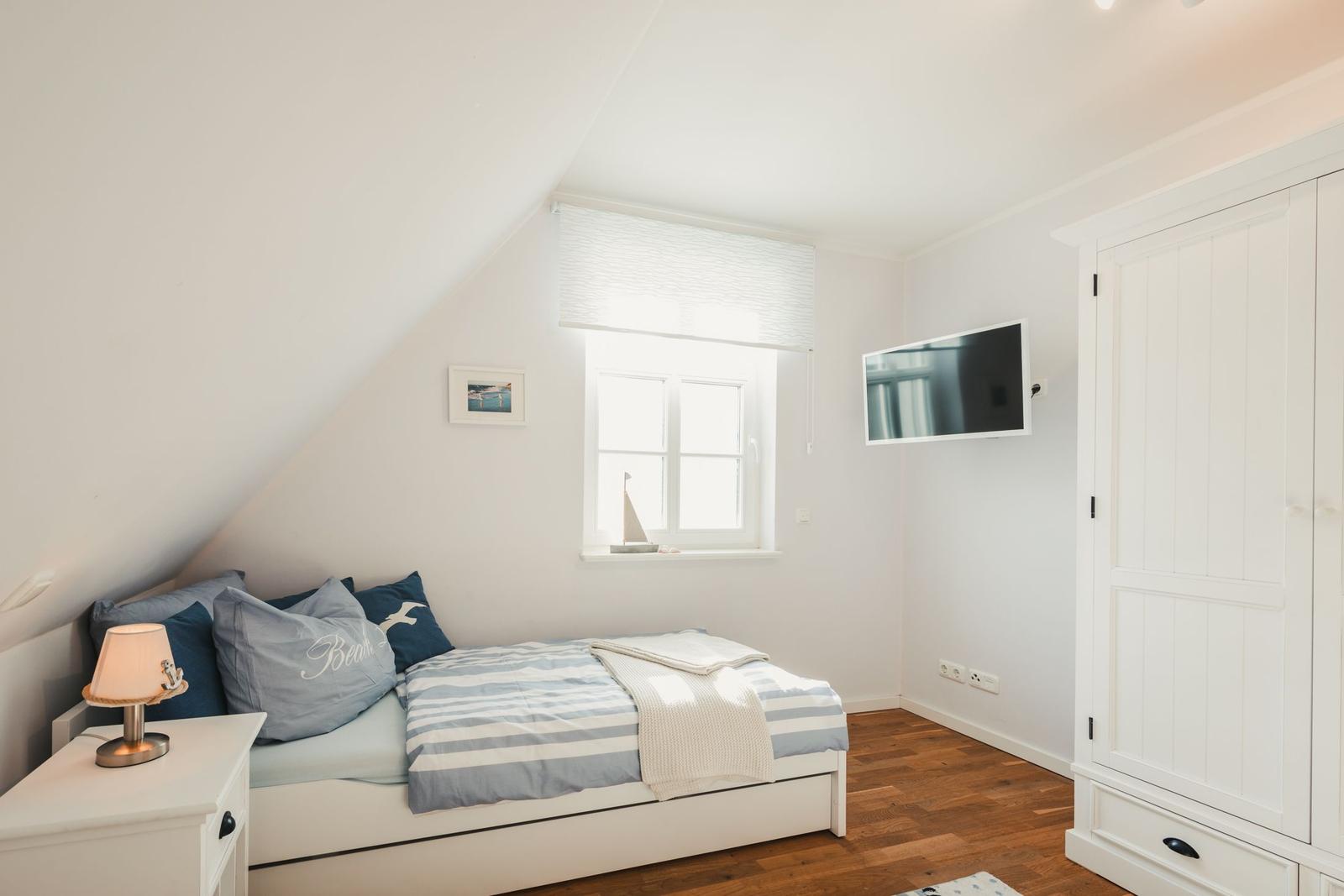 Schlafzimmer mit Bett, Nachttisch, TV und Kleiderschrank. Fenster mit Jalousie und Segelboot auf dem Fensterbrett.