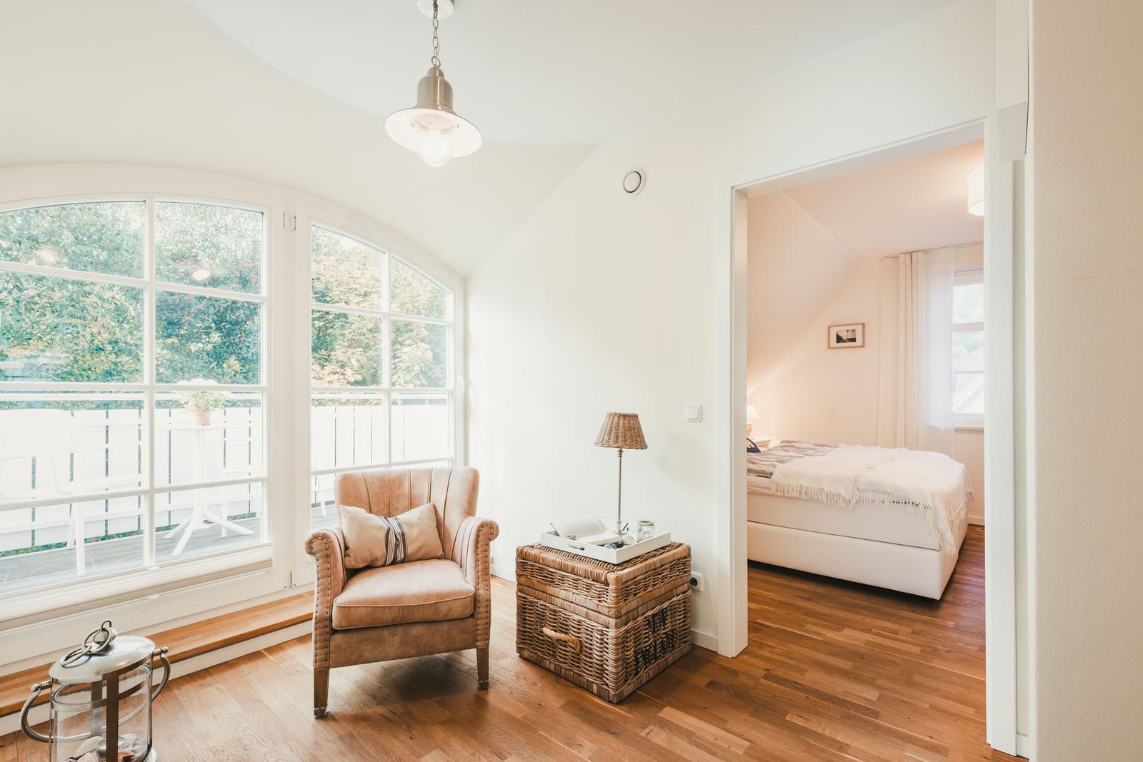 Ein Raum mit Sessel, Korb und großem Fenster. Durchgang zu Schlafzimmer mit Bett.