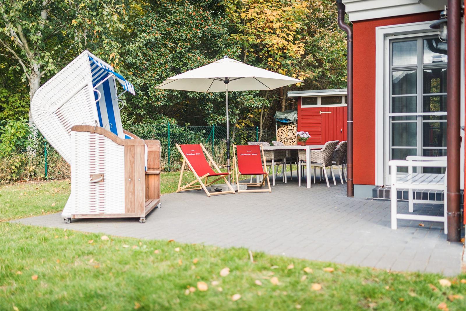 Terrasse mit Liegestühlen, Sonnenschirm und Grillplatz neben rotem Haus.