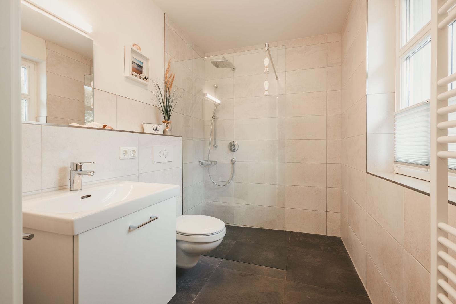 Badezimmer mit Dusche, Waschbecken und Toilette. Fenster und Heizkörper sichtbar.
