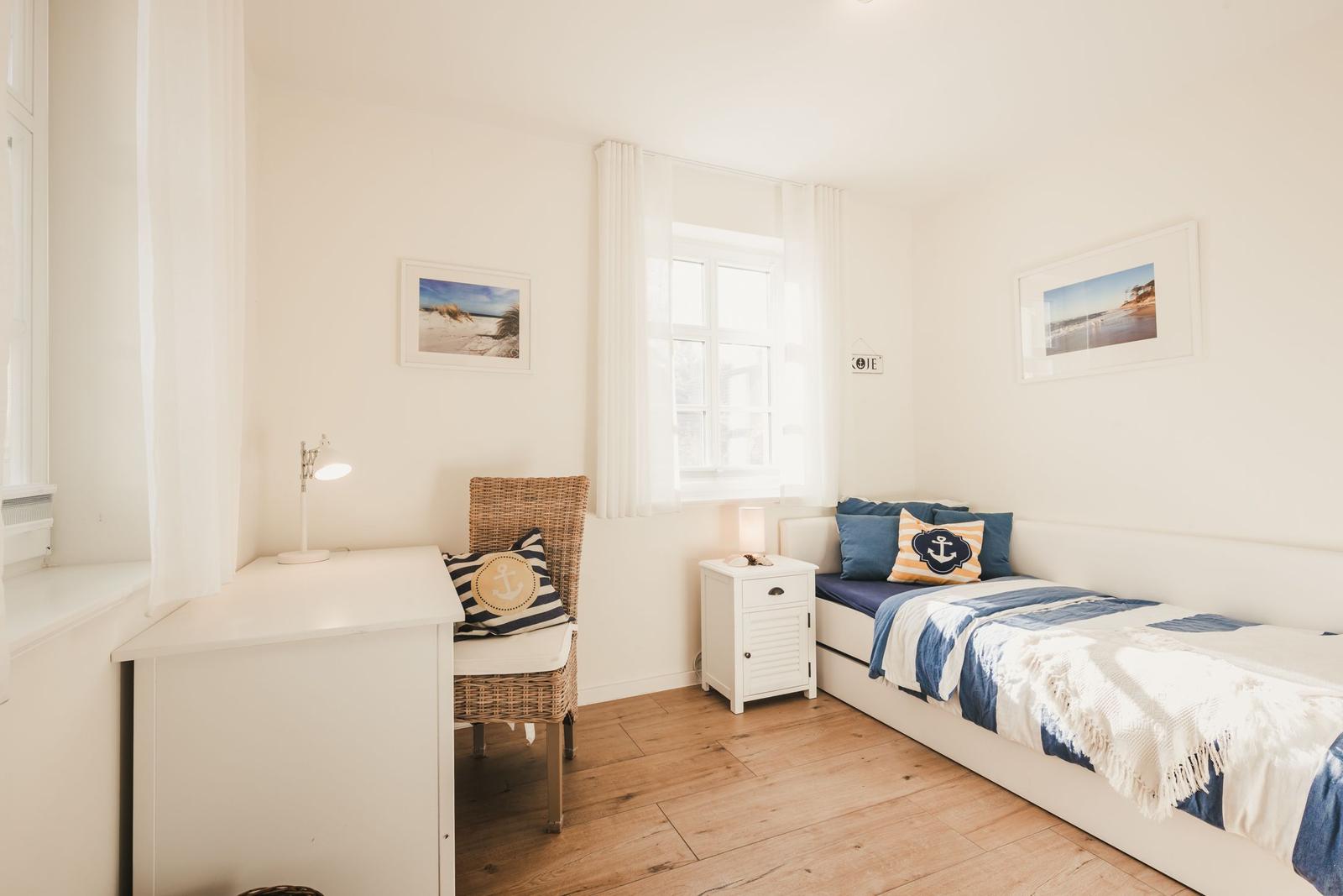 Schlafzimmer mit Bett, Schreibtisch und Fenster. Wandbilder und blaue Kissen.