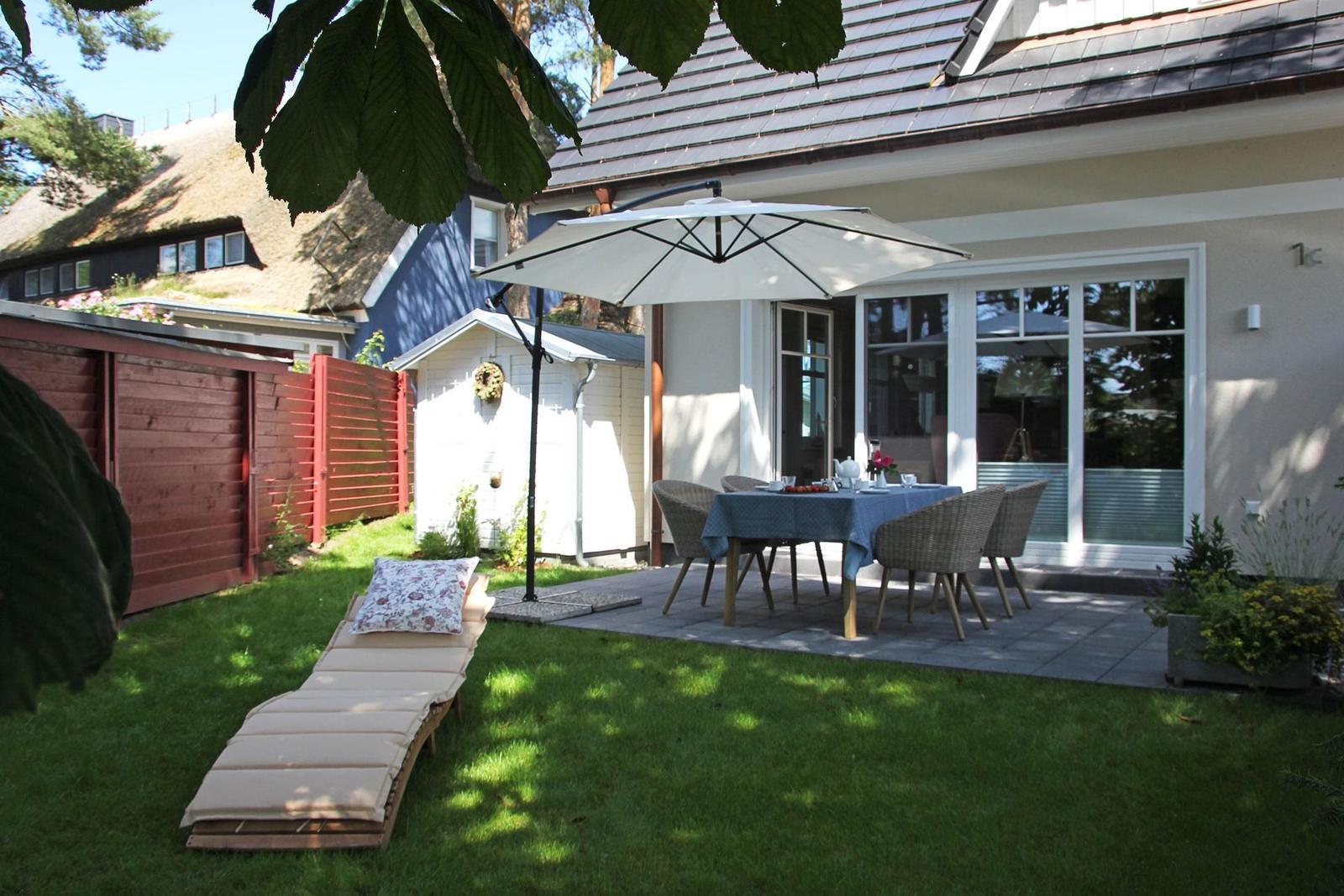 Terrasse mit Tisch, Stühlen und Sonnenschirm. Liegestuhl auf Rasen. Hinterhof mit rotem Zaun.