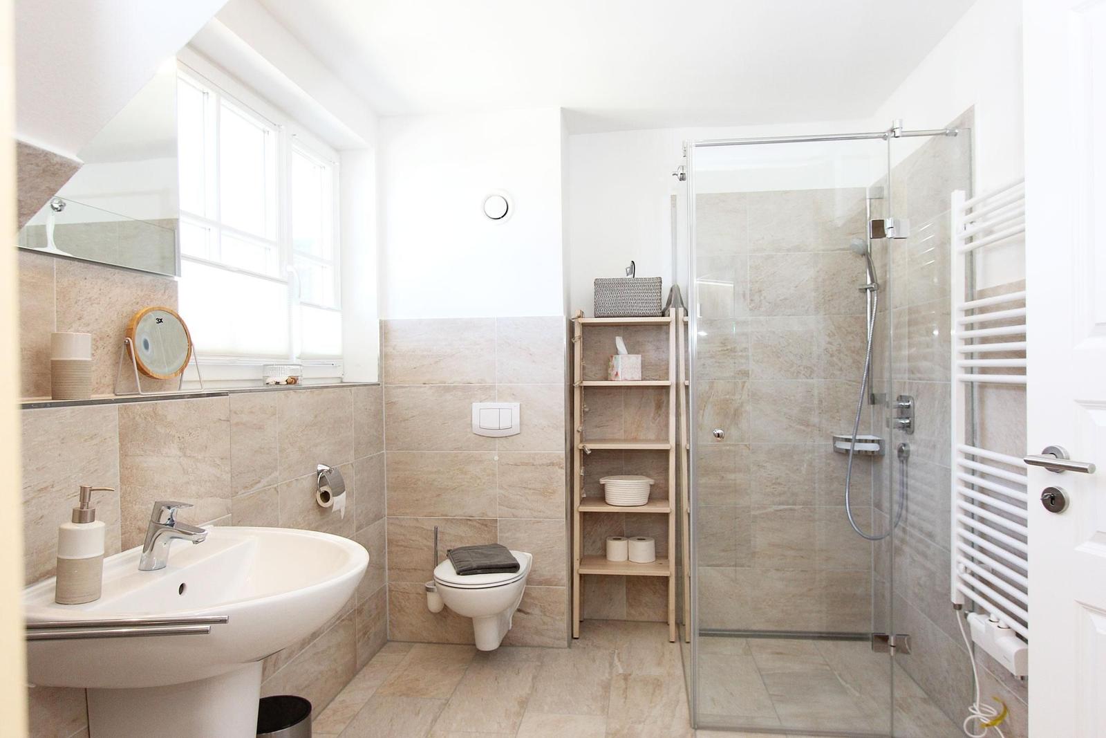 Badezimmer mit Dusche, Toilette und Waschbecken. Holzregal und Heizung sind vorhanden.