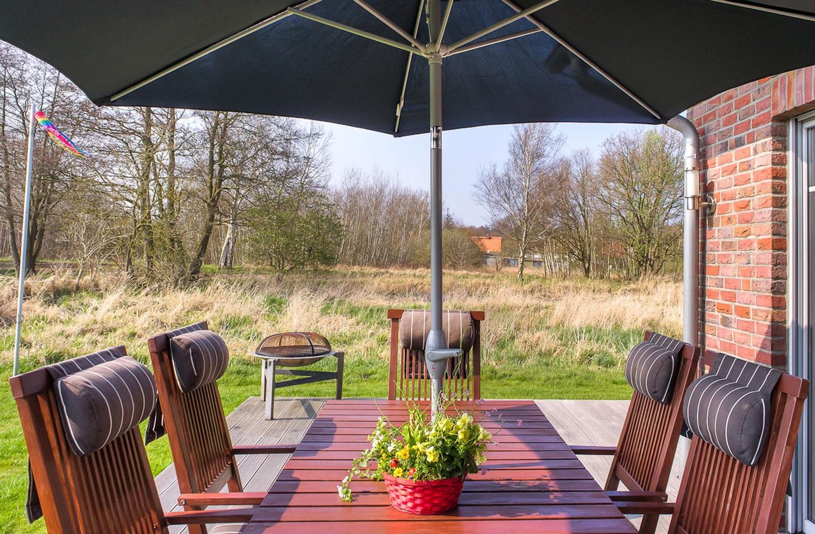 Terrasse mit Tisch, Stühlen und Sonnenschirm. Hintergrund: Grasland und Bäume.