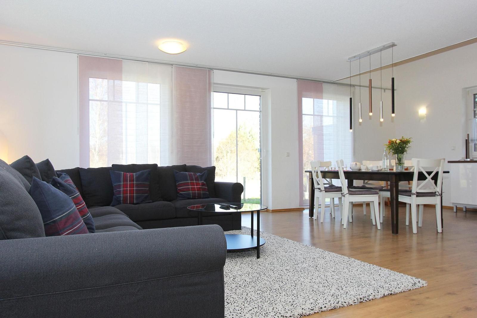 Wohn- und Essbereich mit Sofa, Tisch und Stühlen. Holzboden und große Fenster.
