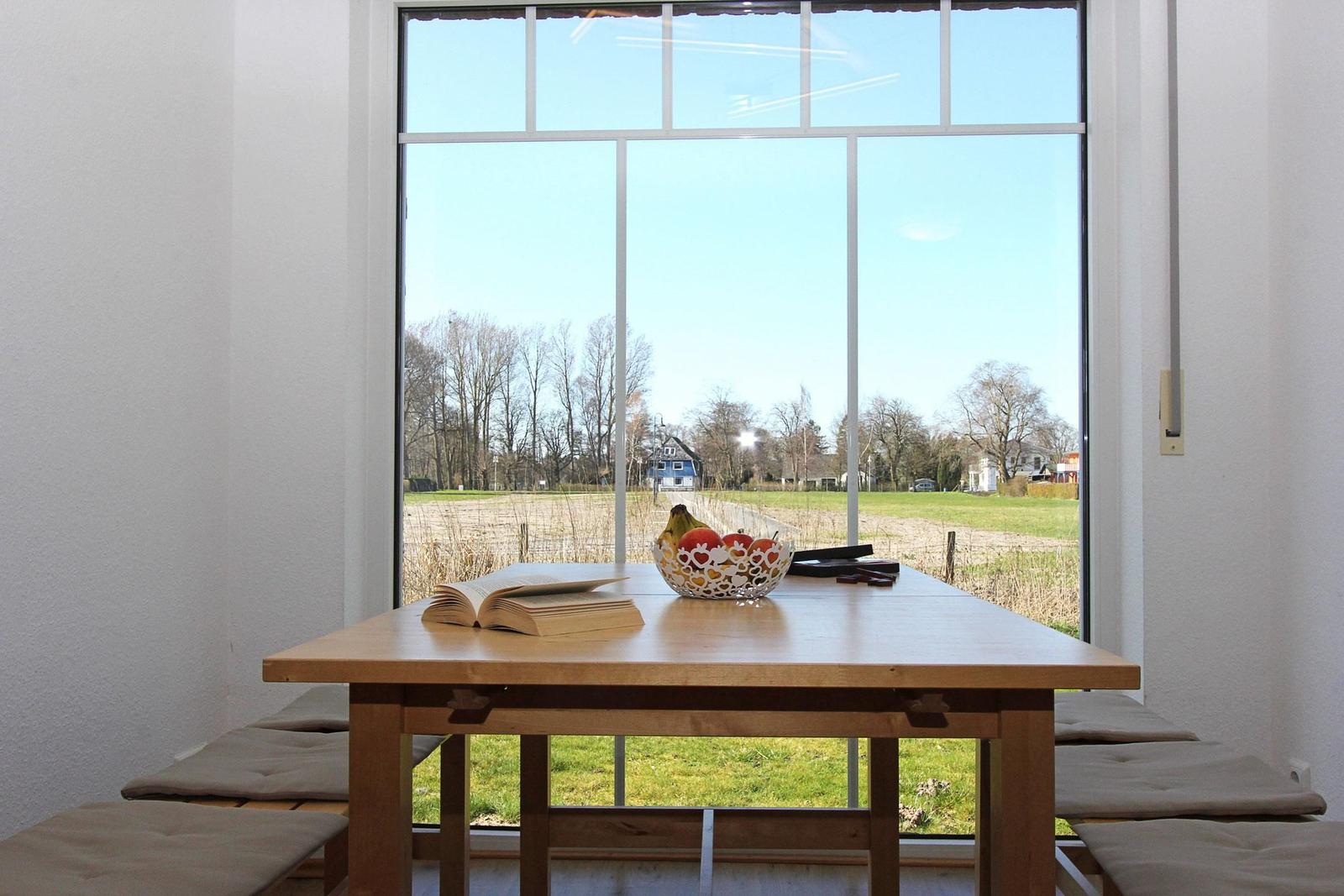 Holztisch mit Obstschale und Buch vor großem Fenster mit Blick auf Feld und Bäume.