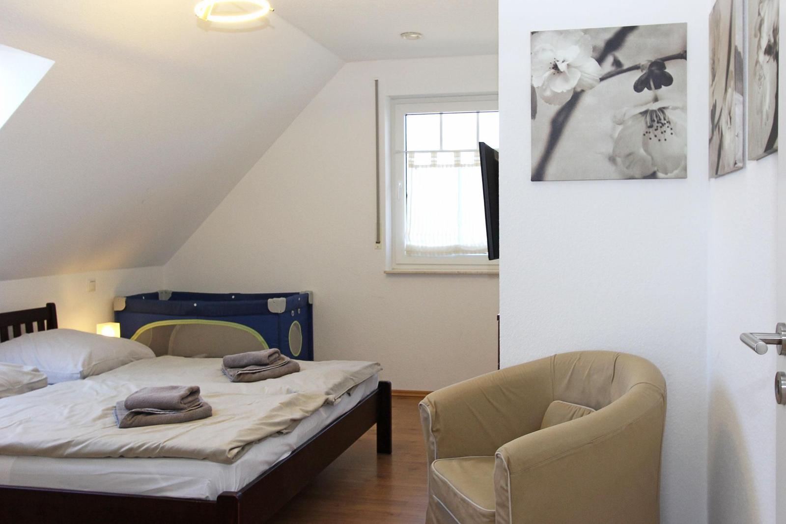 Schlafzimmer mit Bett, Sessel und Fenster. An der Wand hängt ein Bild mit Blumen.