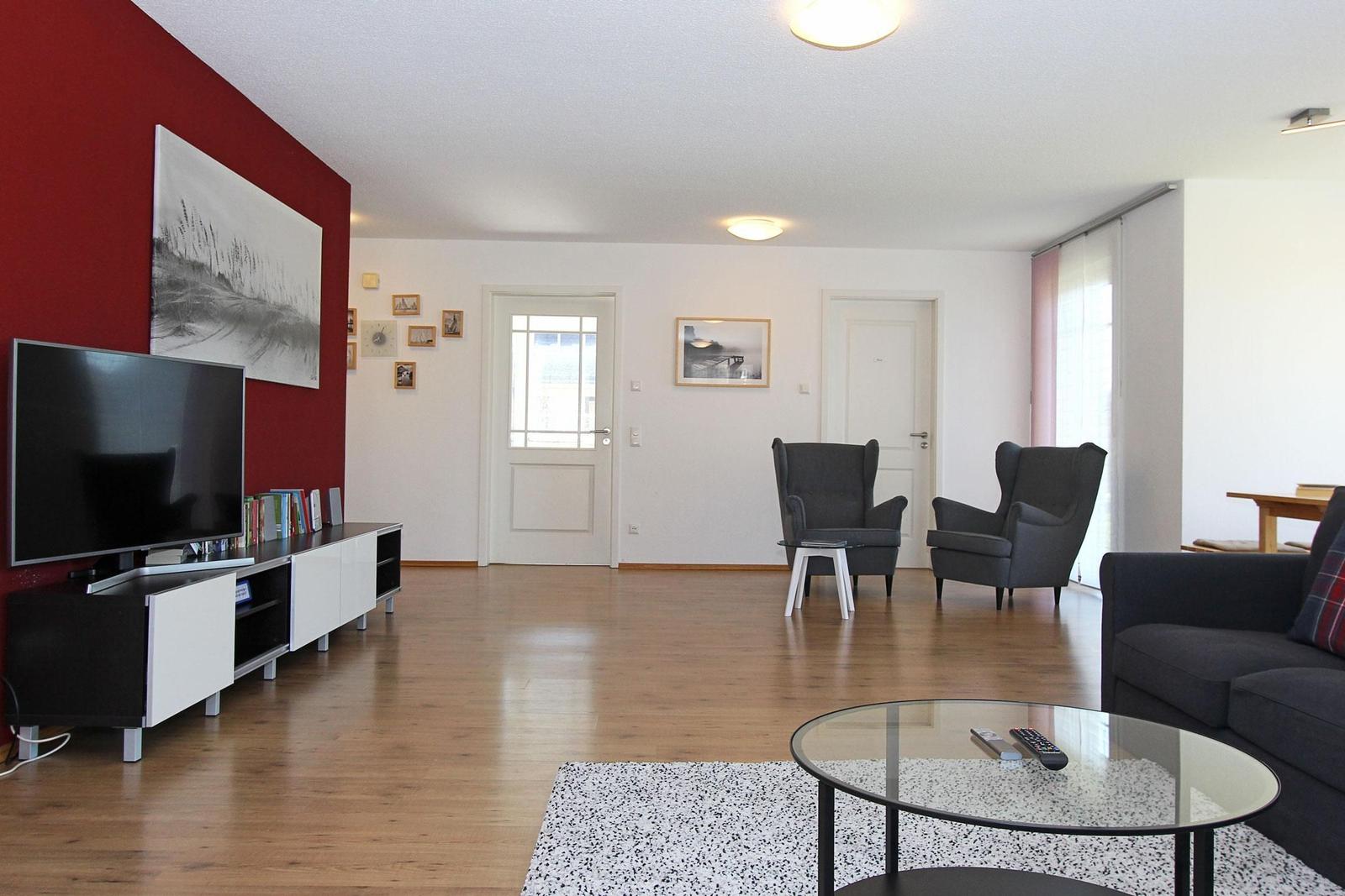 Wohnzimmer mit Holzboden, Sofa, zwei Sessel und Fernseher an roter Wand.