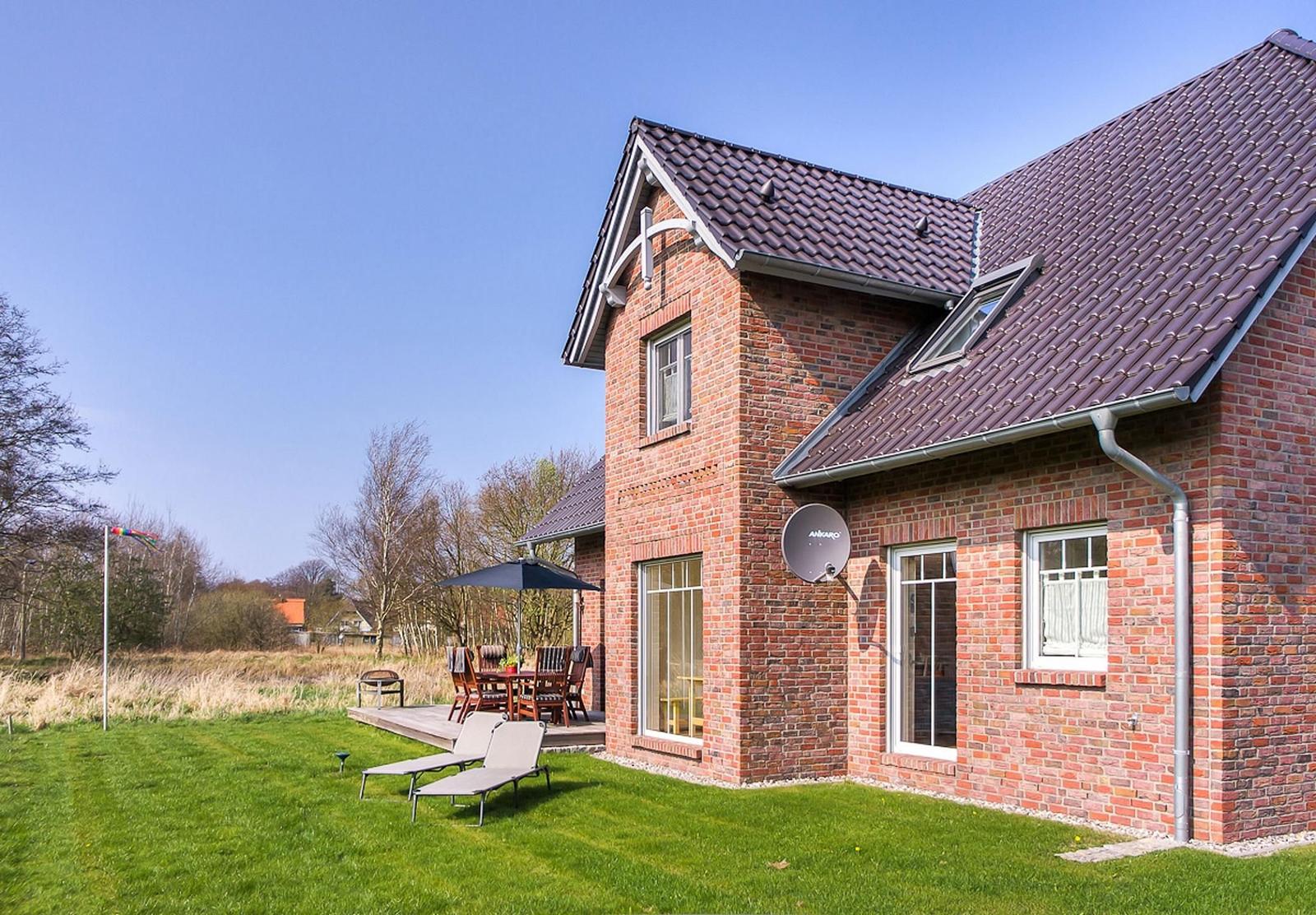 Rotes Backsteingebäude mit Terrasse, Liegestühlen und Satellitenschüssel. Grünflächen und Bäume im Hintergrund.