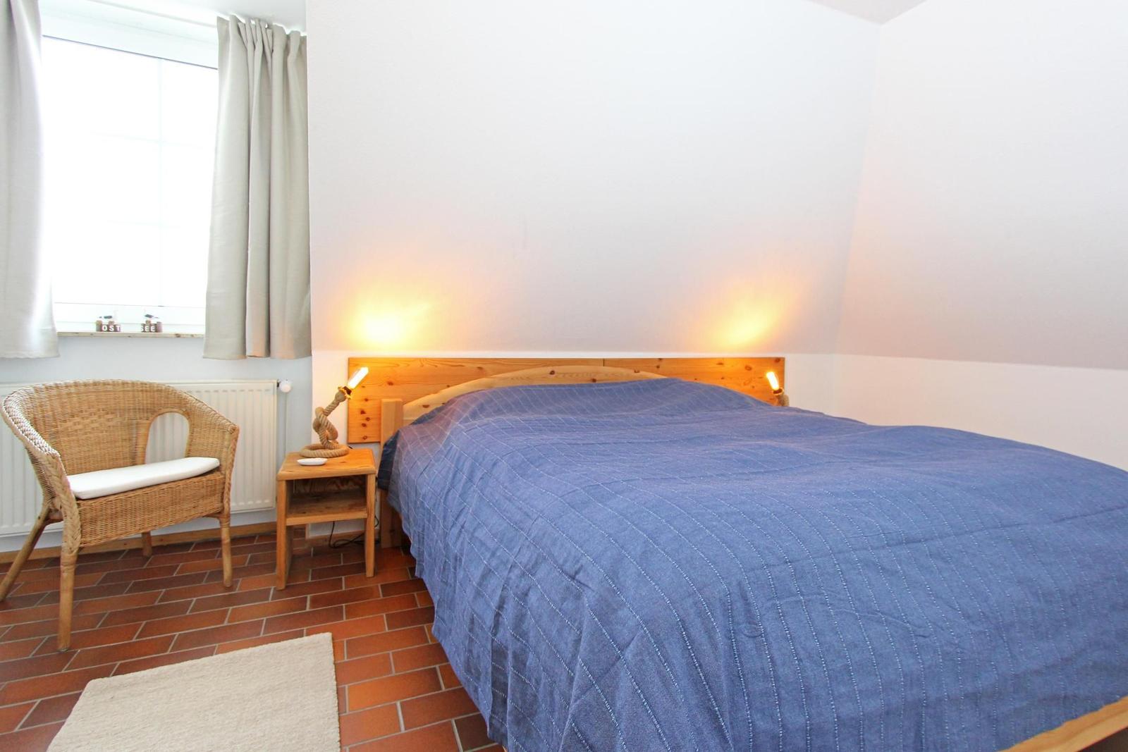 Schlafzimmer mit Bett, Nachttisch und Stuhl. Fenster mit Vorhängen und Bodenfliesen.