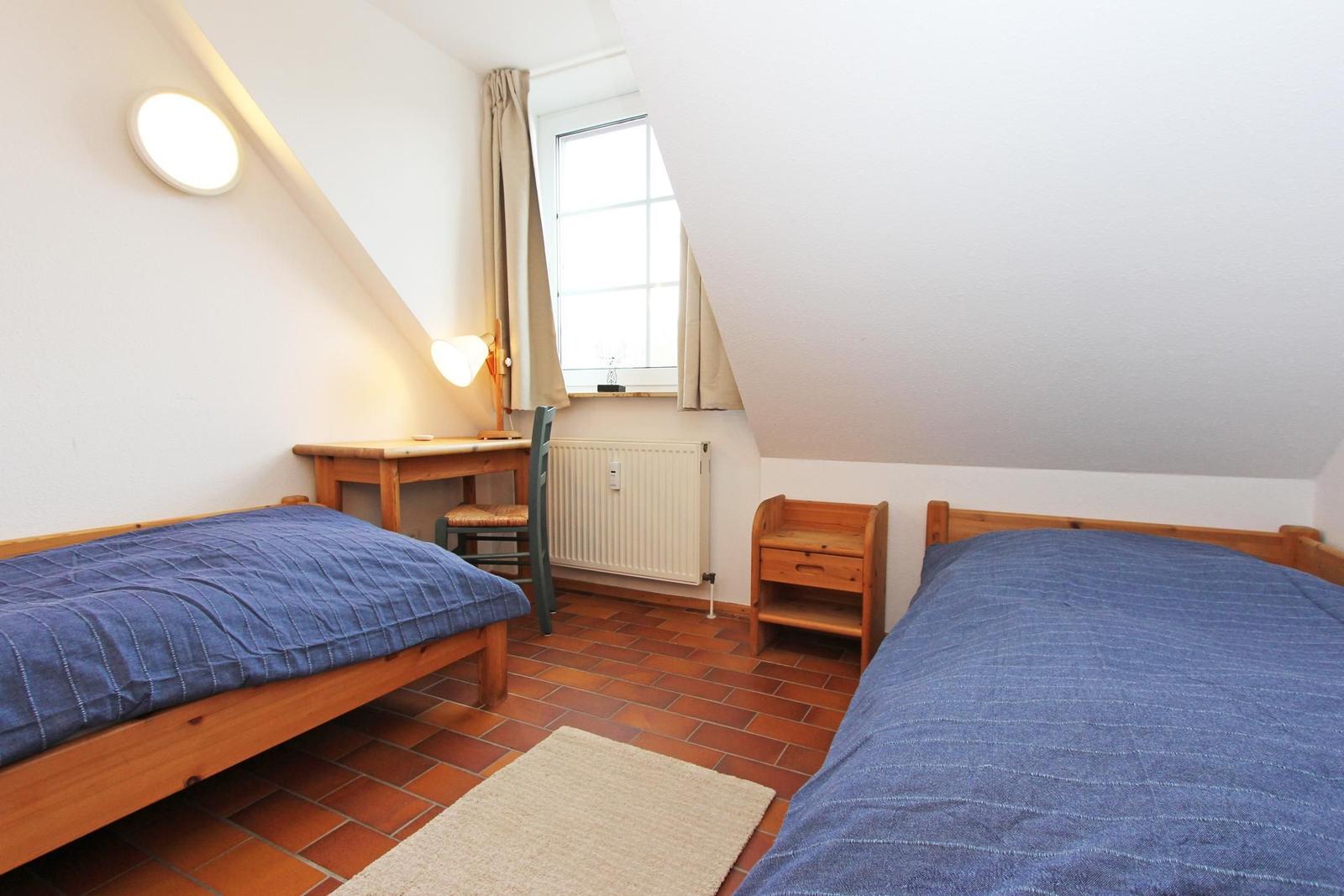 Zwei Betten, Schreibtisch, Fenster und Holzmöbel in einem Dachzimmer mit Terrakottboden.