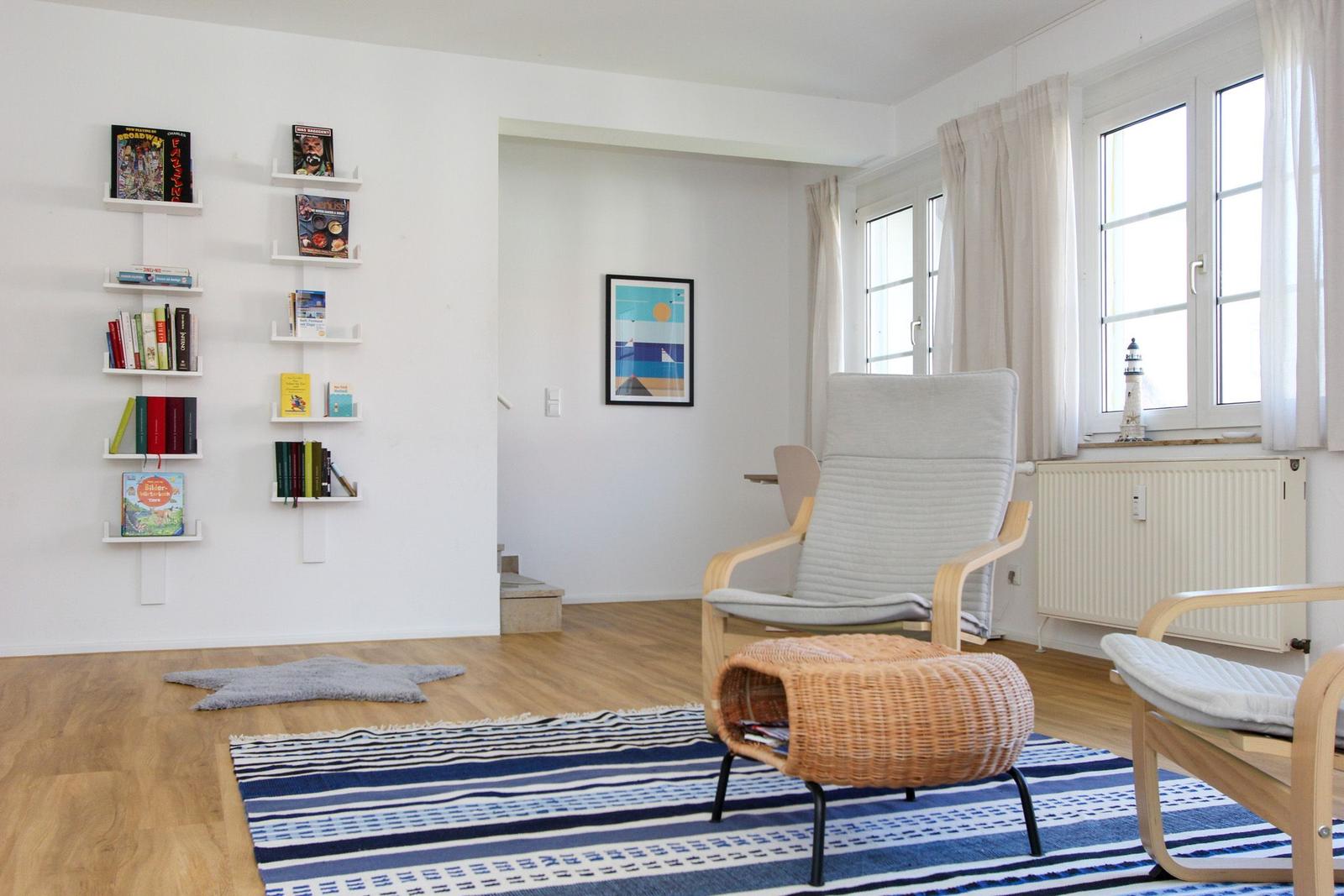 Wohnzimmer mit Bücherregal, Sessel und Fenster. Teppich und Kissen auf Holzboden.
