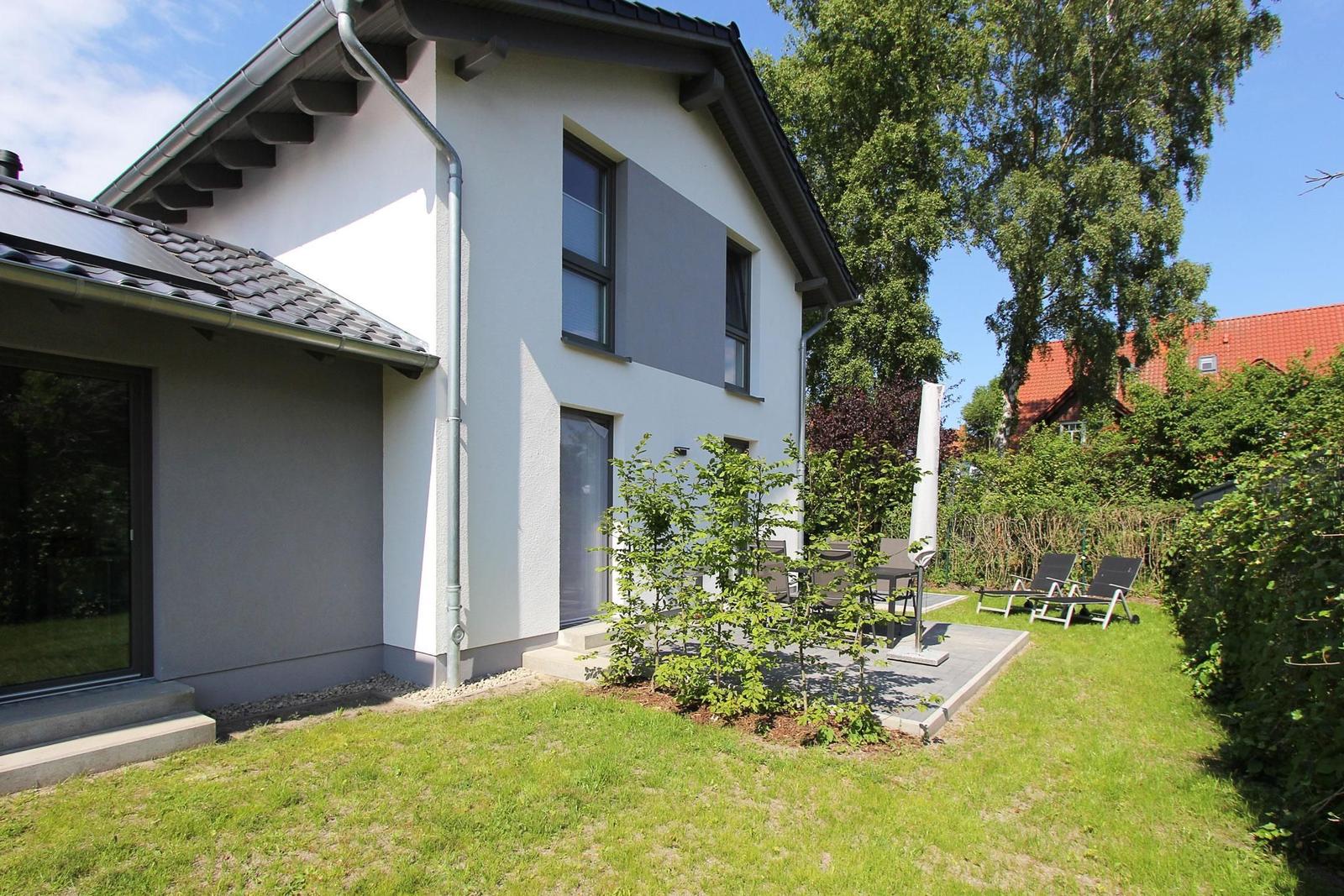 Moderne Villa mit Garten, Terrasse und Liegestühlen. Schwarze Fenster und graue Fassade.