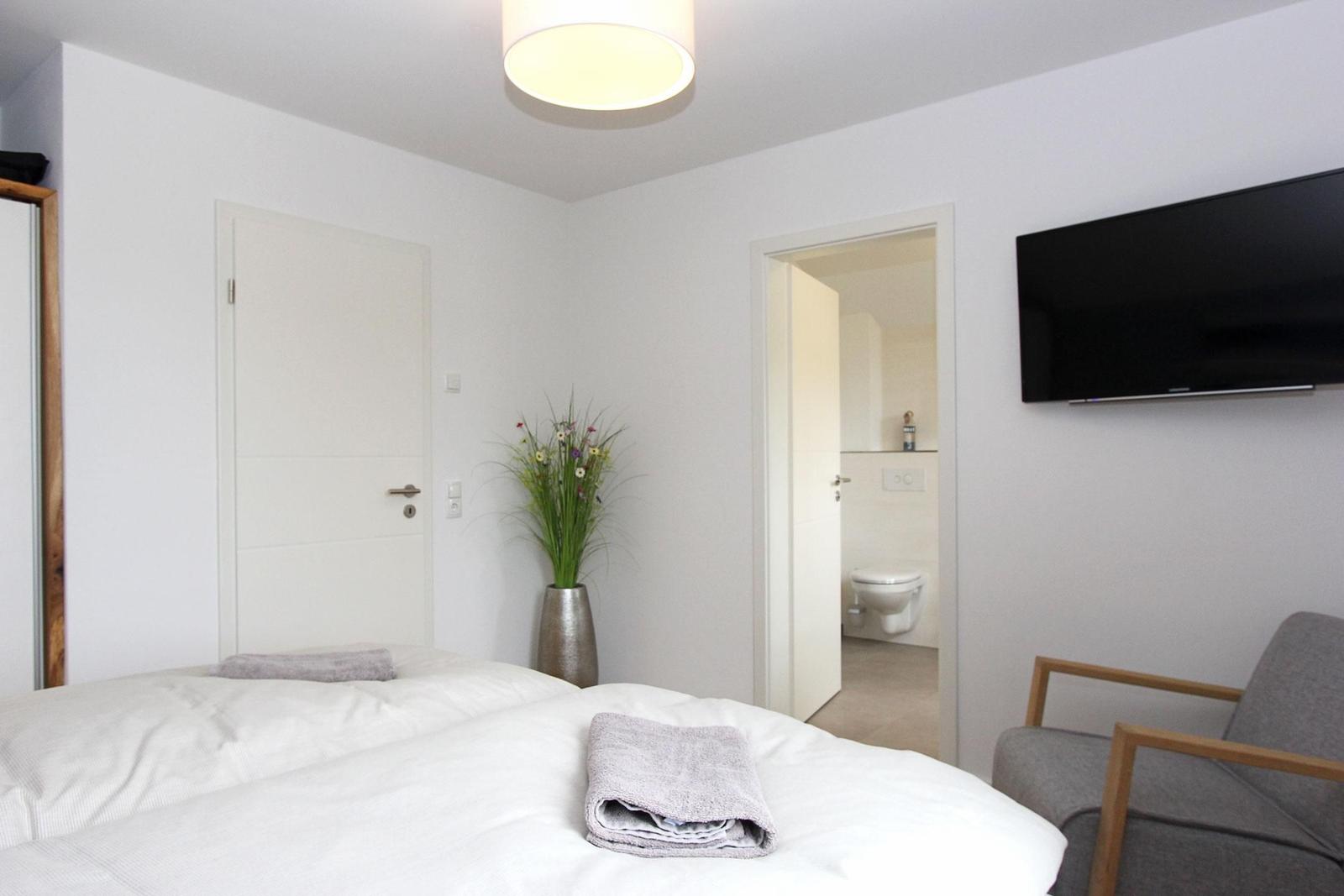 Schlafzimmer mit Bett, Fernseher und Sessel. Flur führt in die Toilette.