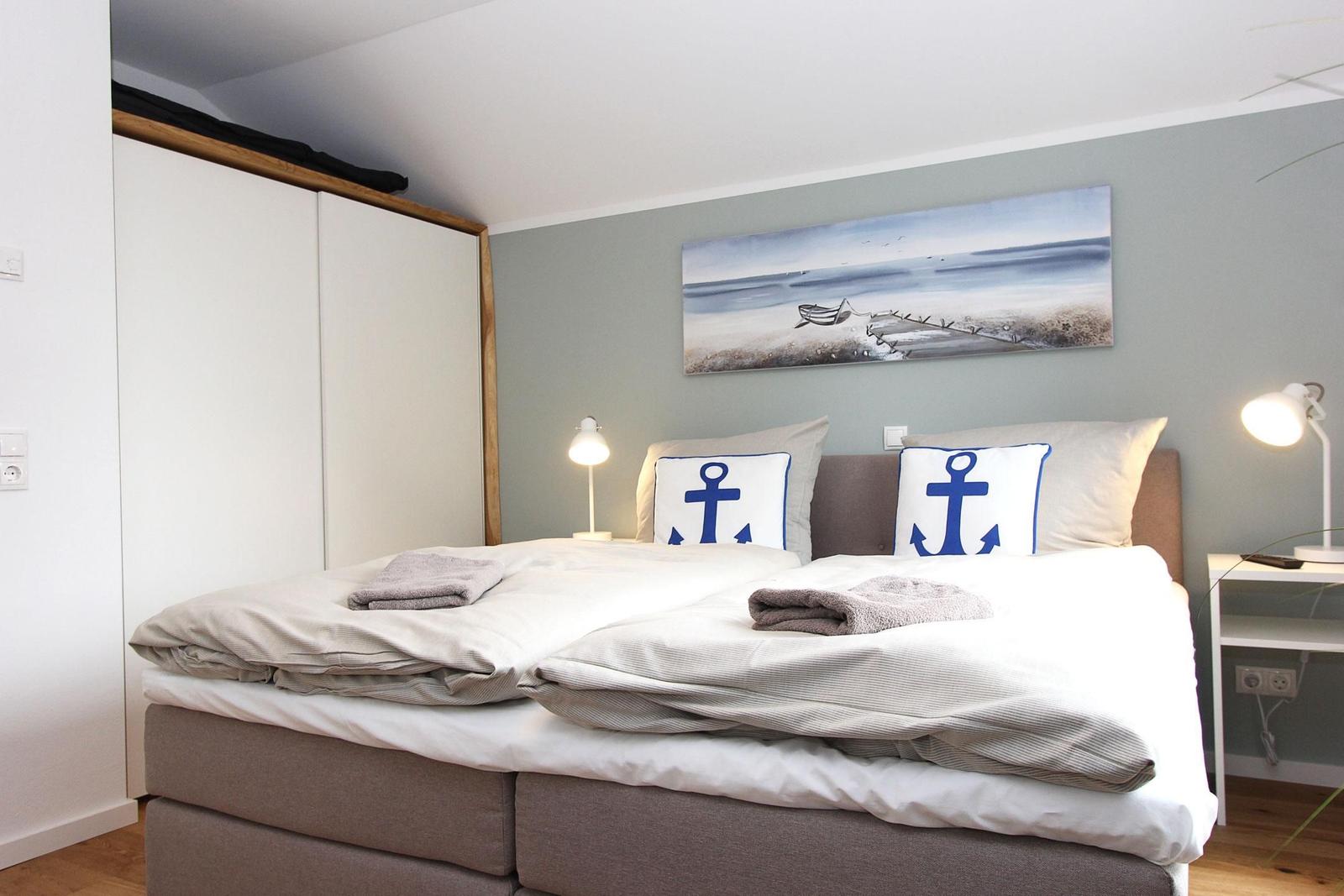 Schlafzimmer mit Doppelbett, Ankerkissen und Wandbild. Links Schrank, rechts Nachttisch mit Lampe.
