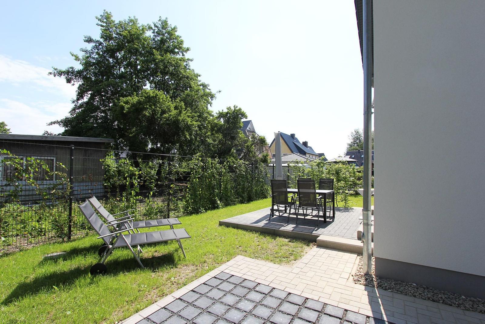Hinterhof mit Liegestühlen und Tischset auf Terrasse. Grünfläche und Zaun sichtbar.