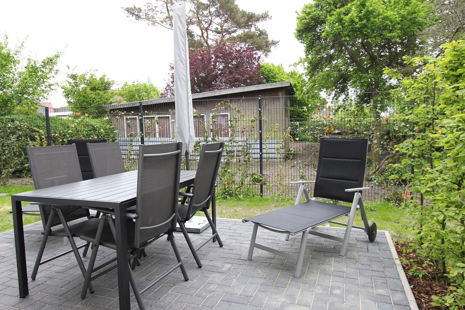 Terrasse mit Tisch, Stühlen und Liegestuhl. Hintergrund: Zaun und Haus.