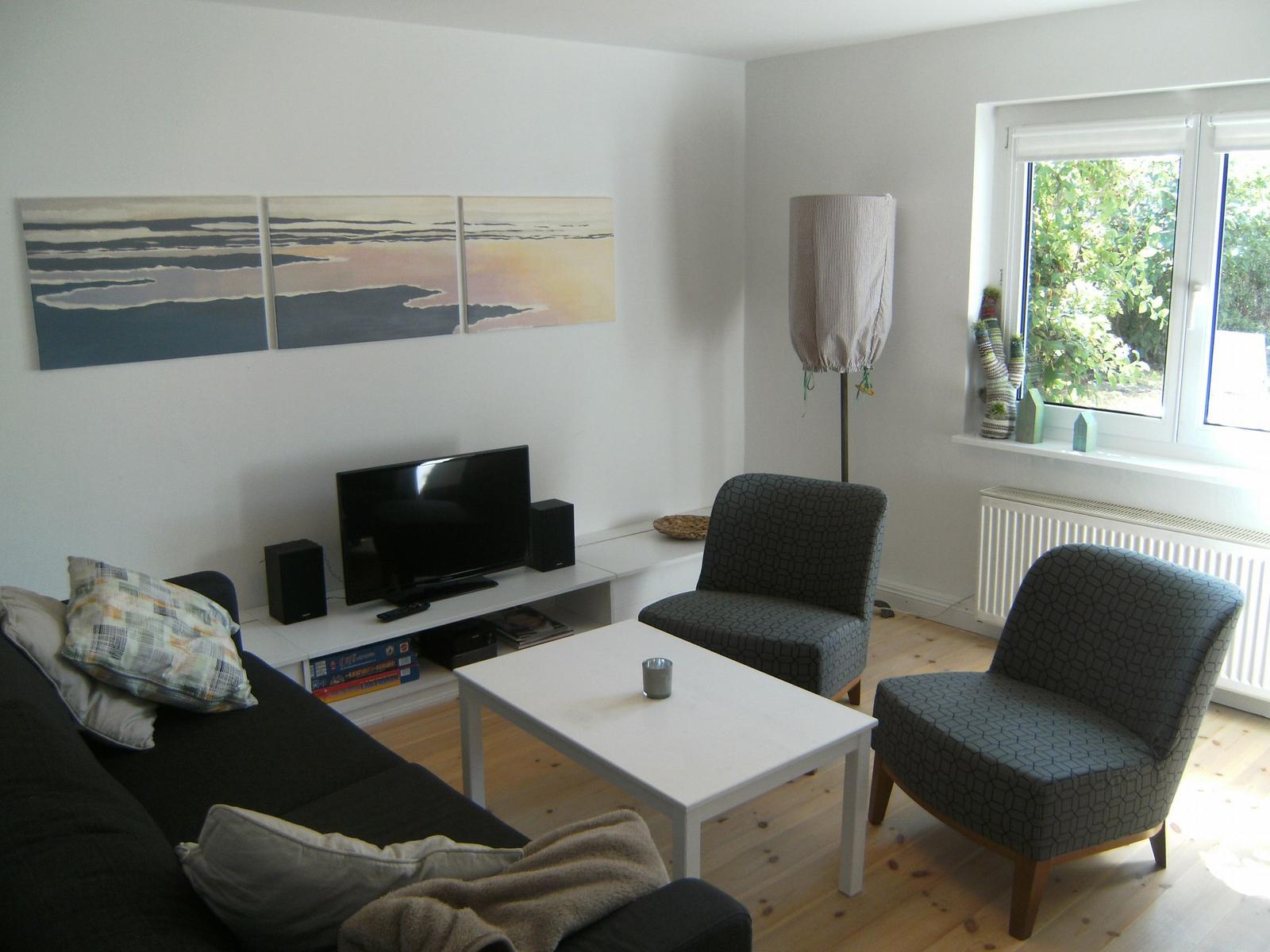 Wohnzimmer mit Sofa, zwei Sesseln, TV und Fenster mit Blick auf Garten.