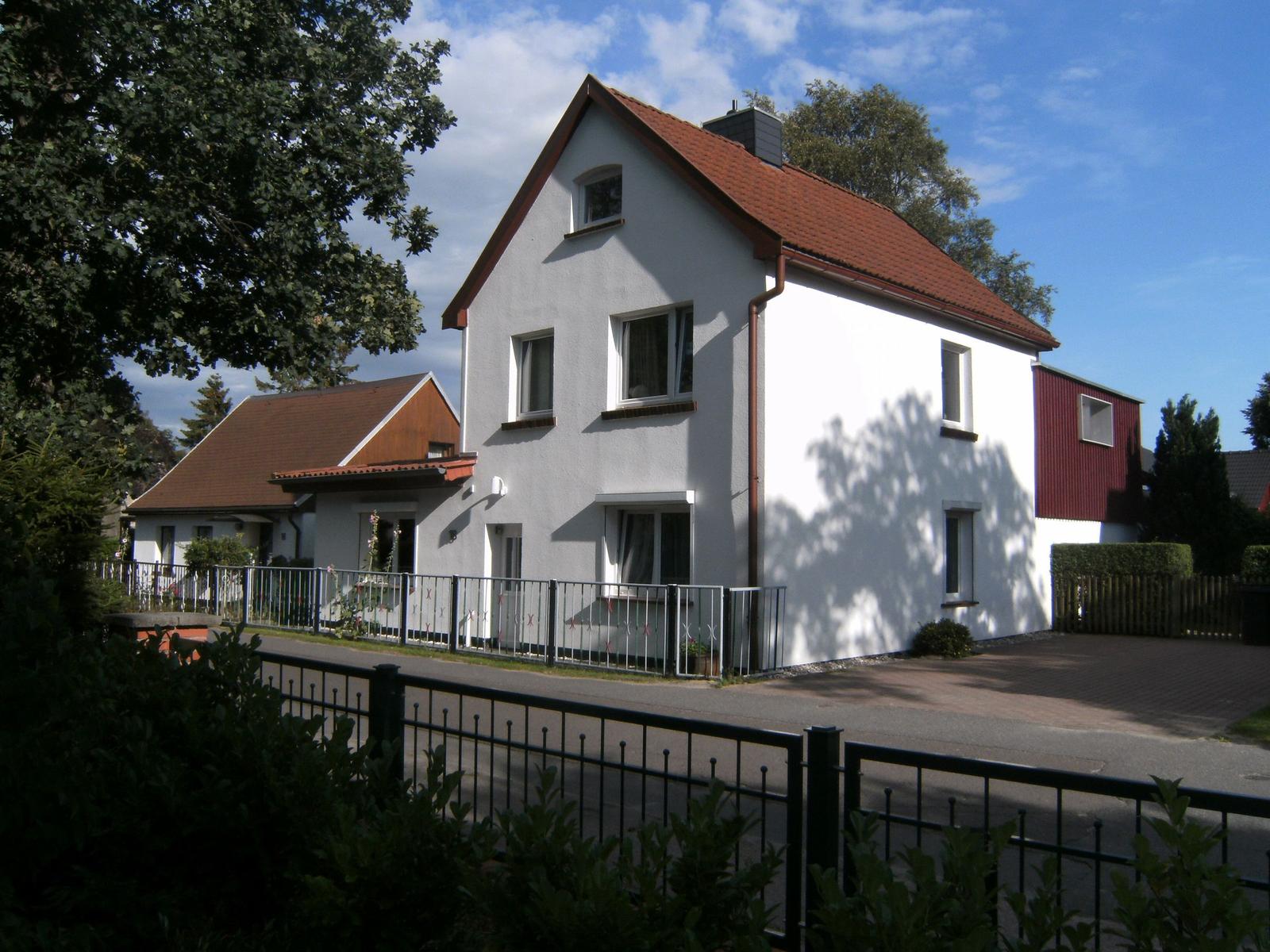 Weiße Villa mit rotem Dach und Metallzaun. Umgeben von Bäumen und Grünflächen.