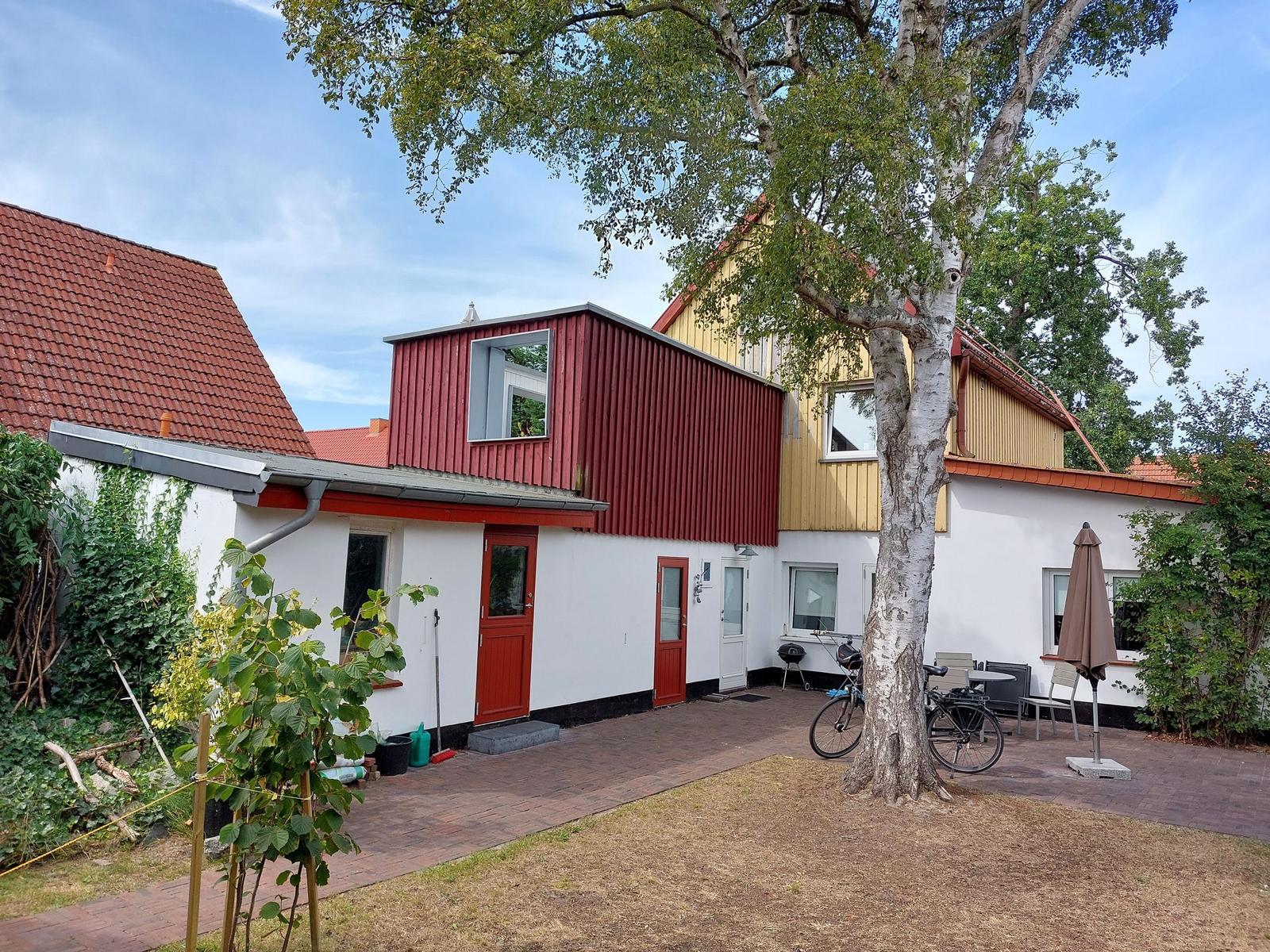 Ein Haus mit rotem und gelbem Anbau, Terrasse mit Tisch, Stühlen und einem Baum.