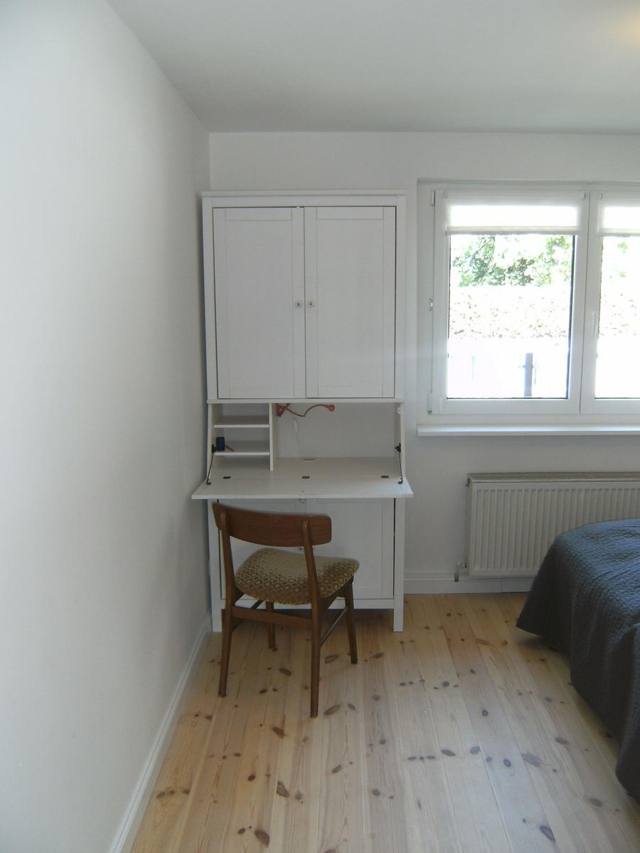 Schlafzimmer mit Schreibtisch, Stuhl und Fenster. Holzfußboden und Bett mit grauer Bettwäsche.