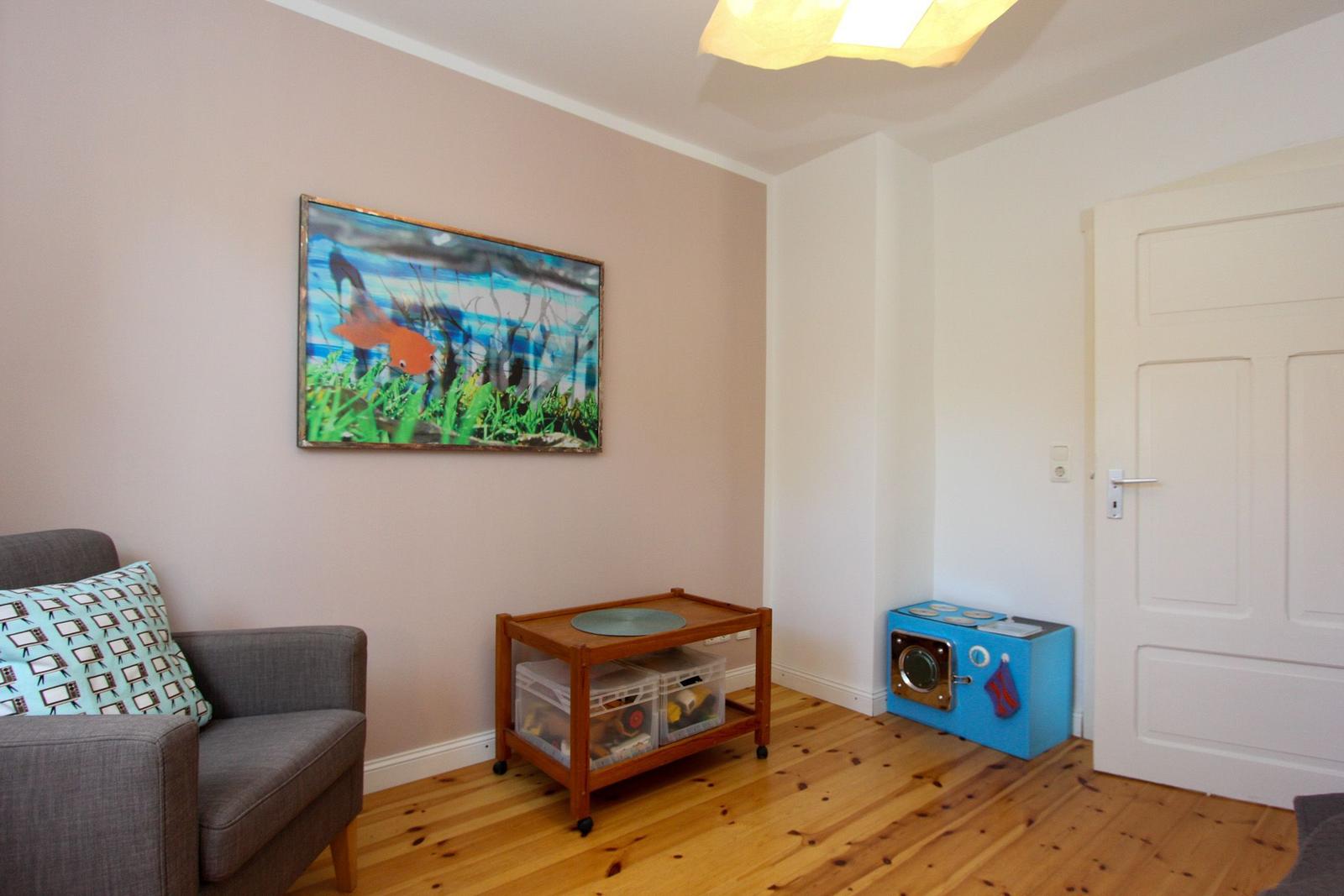 Ein Raum mit grauem Sessel, Holzboden und Wandbild mit Fisch. Ein blauer Kinderkochblock steht an der Tür.