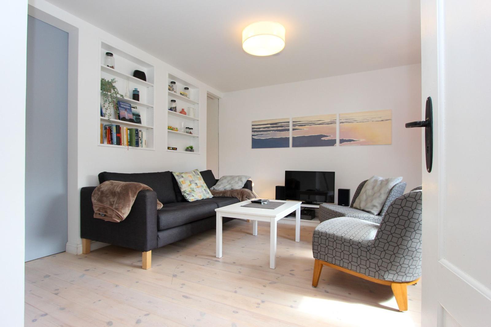Wohnzimmer mit Sofa, Sessel, Couchtisch und Wandregalen. An der Wand hängen drei Bilder.