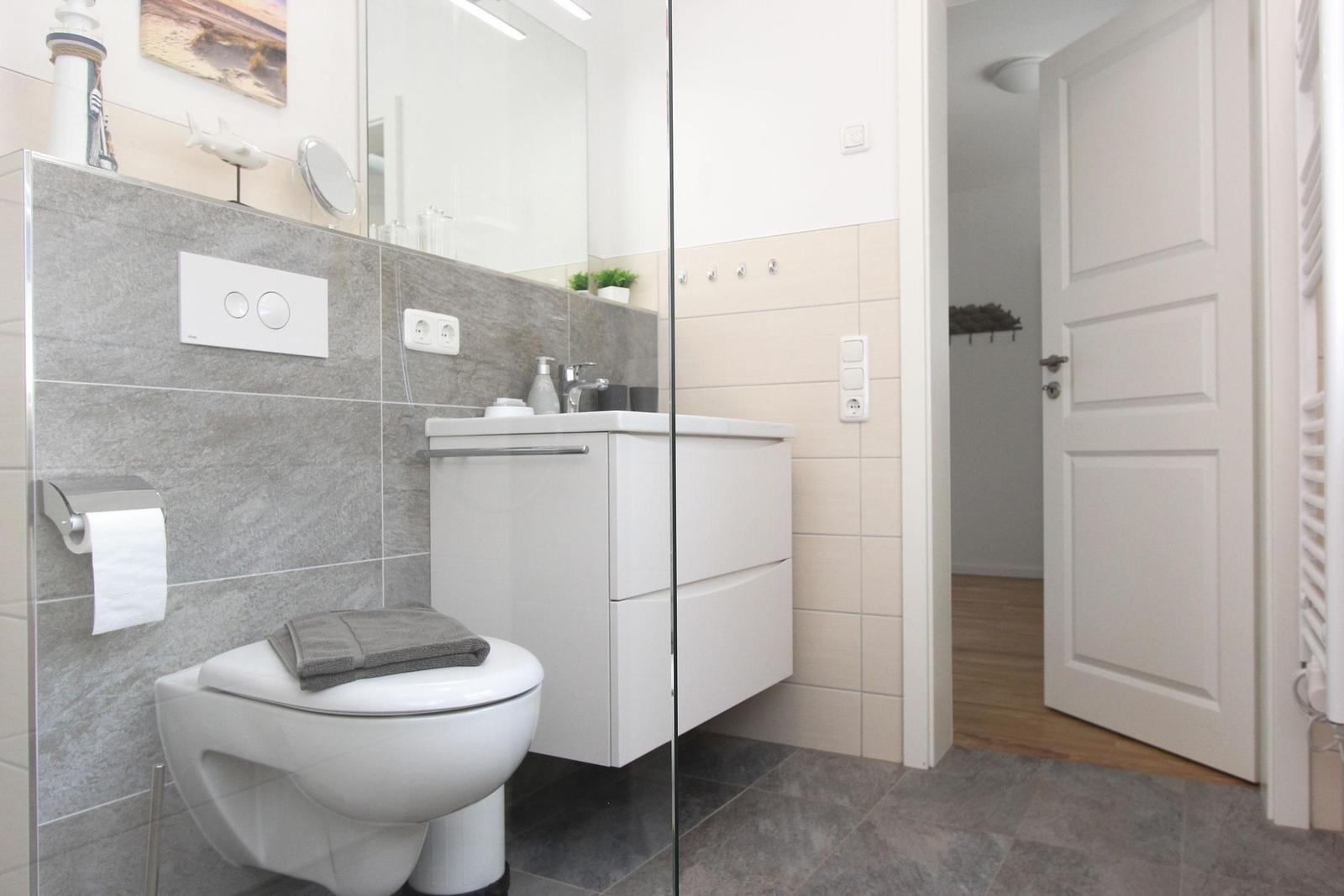 Badezimmer mit Toilette, Waschbecken und Dusche. Graue Fliesen und weiße Möbel.