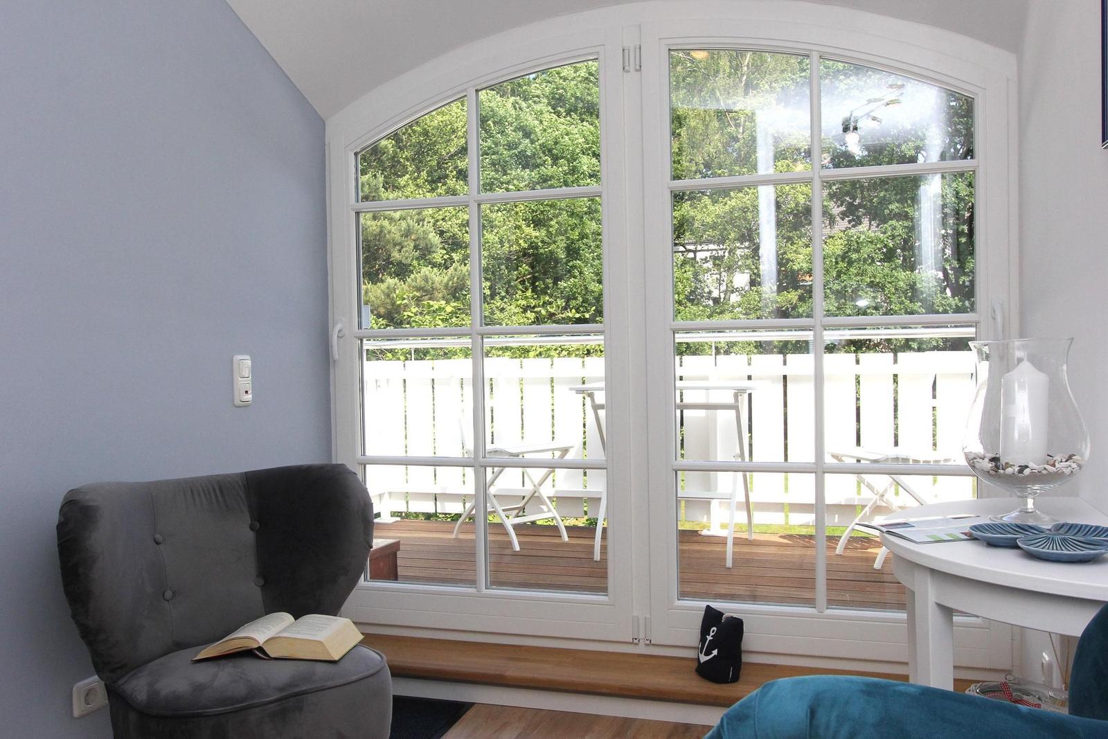 Ein Raum mit grauem Sessel, großem Fenster und Balkon mit Gartenblick.