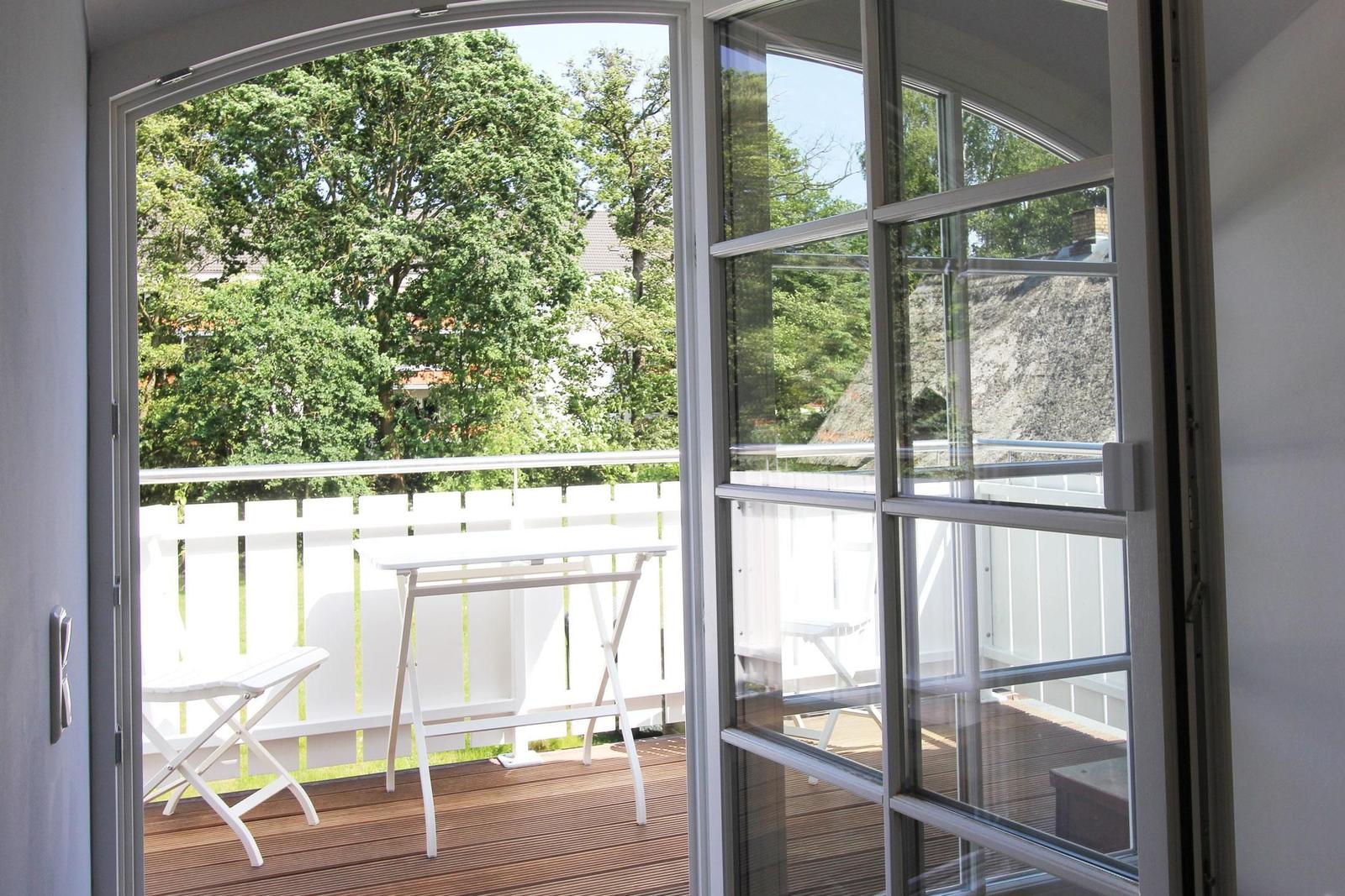 Durch die Glastür ist eine Terrasse mit Tisch und Stühlen zu sehen. Im Hintergrund sind Bäume und ein Dach sichtbar.